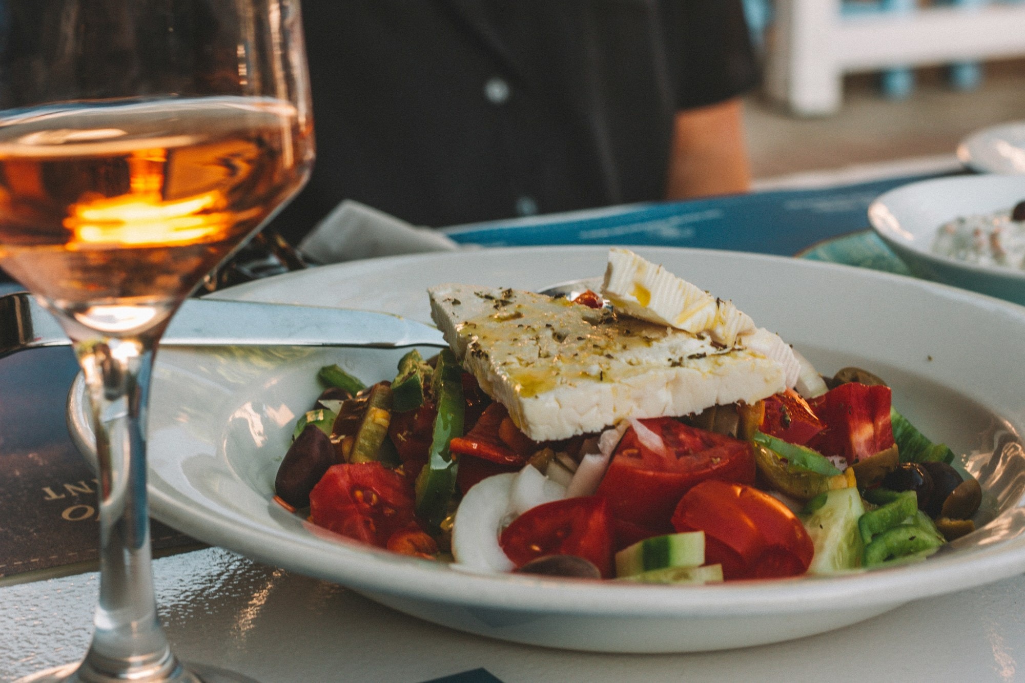 A Greek salad with a block of feta next to a glass of orange wine on a table.