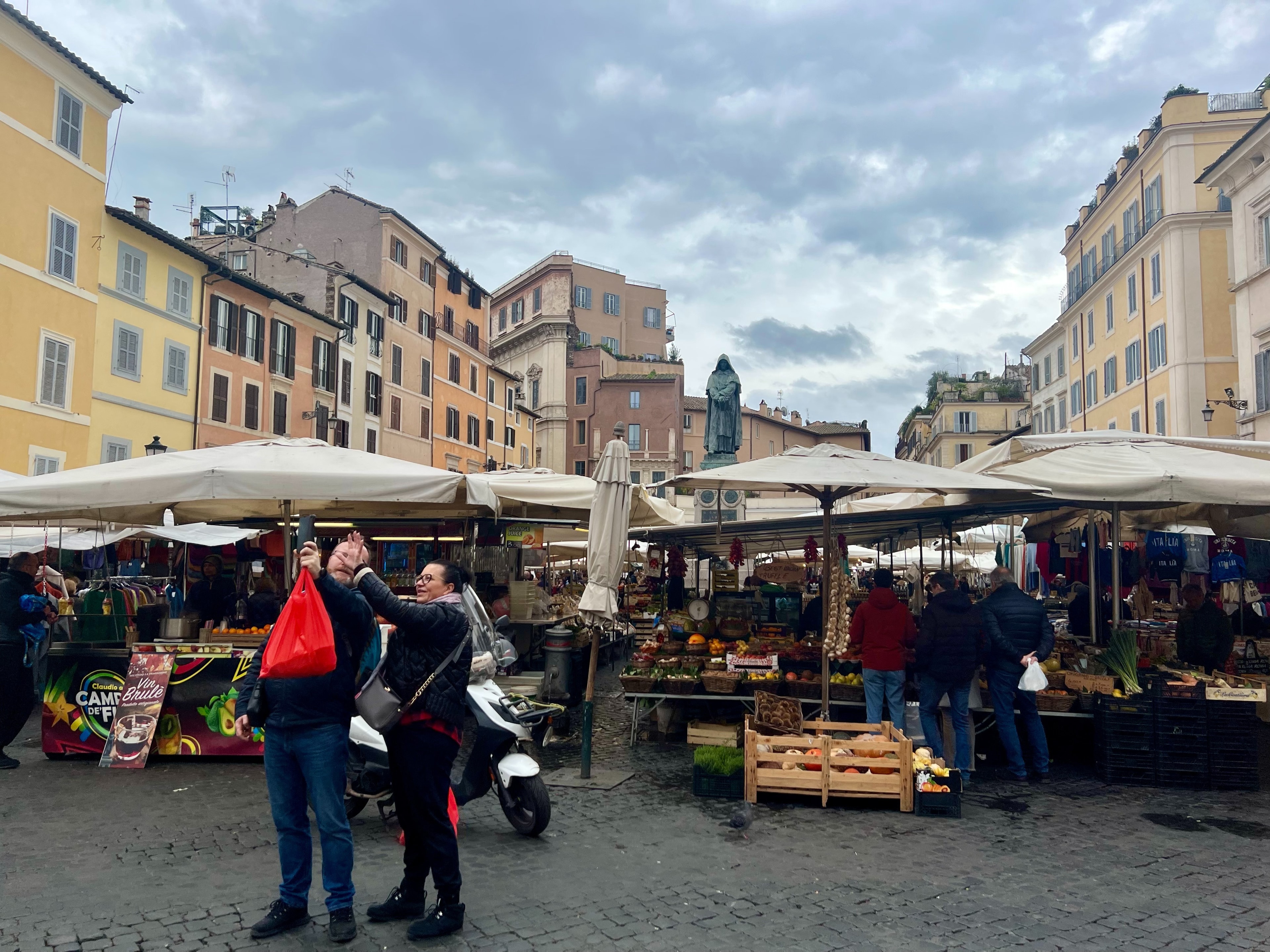 Two tourists taking photos in front of an outdoor market in a city square.