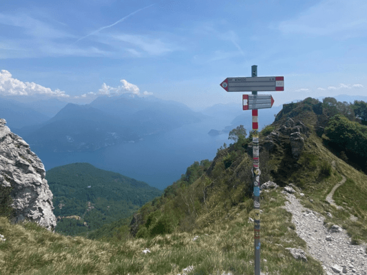 A signpost on a mountain trail, pointing the way against a backdrop of lake and peaks under a clear blue sky.