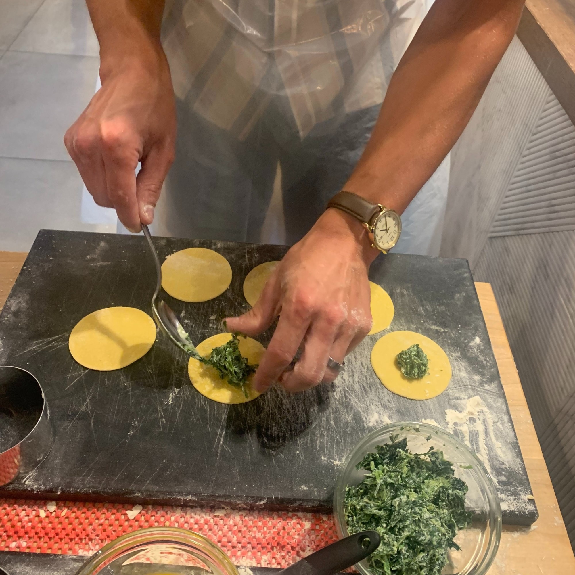 This image depicts a person constructing pasta over a dark board. There are bowls of ingredients in the forefront of the image. 