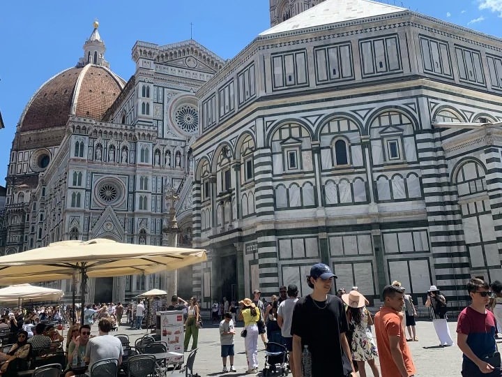 This picture depicts a crowd of tourists walking in front of the duomo in Florence, Italy with it's stone architecture in front of a blue sky. There are dining tables, chairs and yellow umbrellas on the bottom left side of the picture, too. 