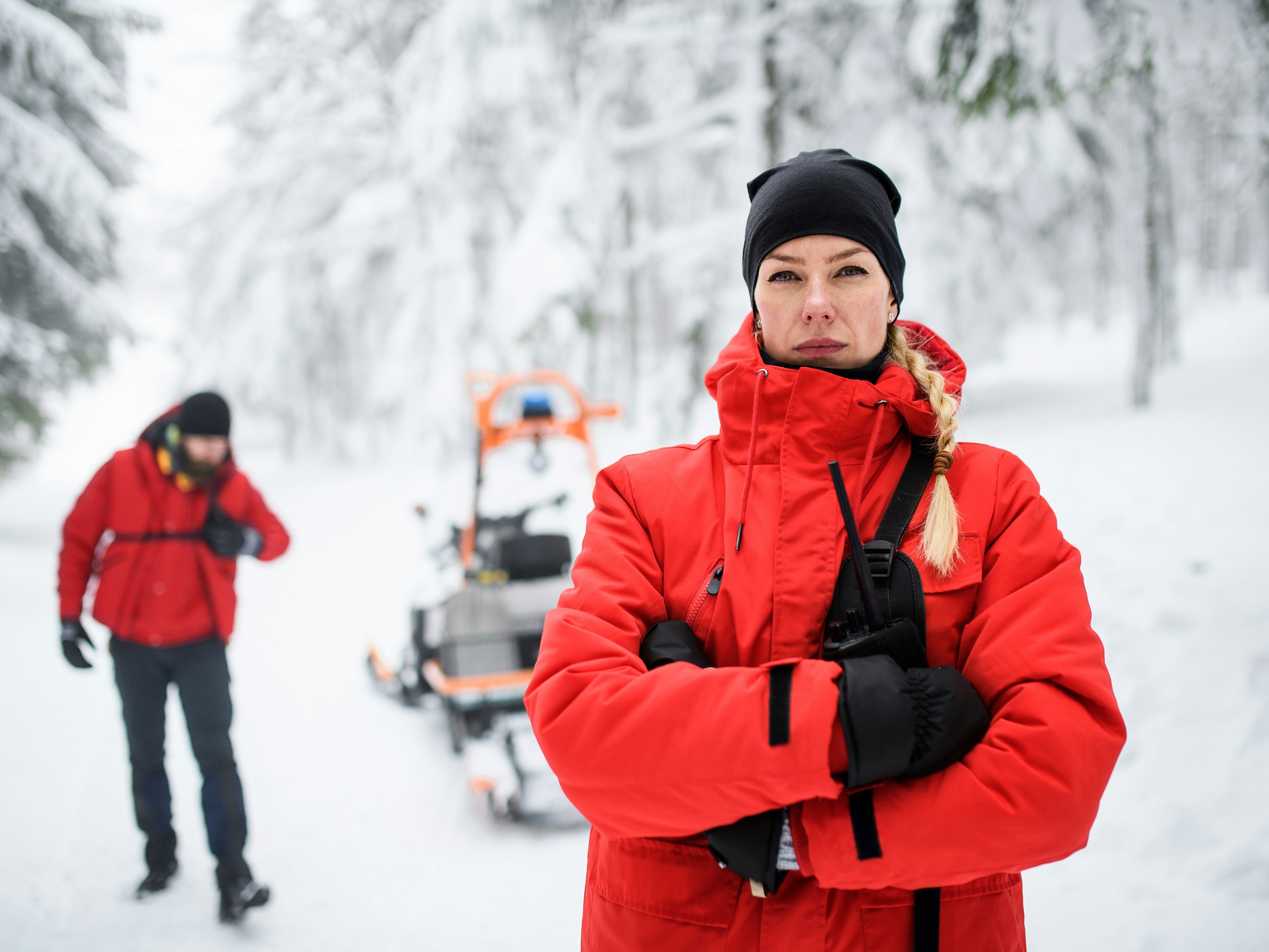 Two individuals and a snowmobile in a snowy forest; one in a red jacket with arms crossed, the other near the snowmobile.