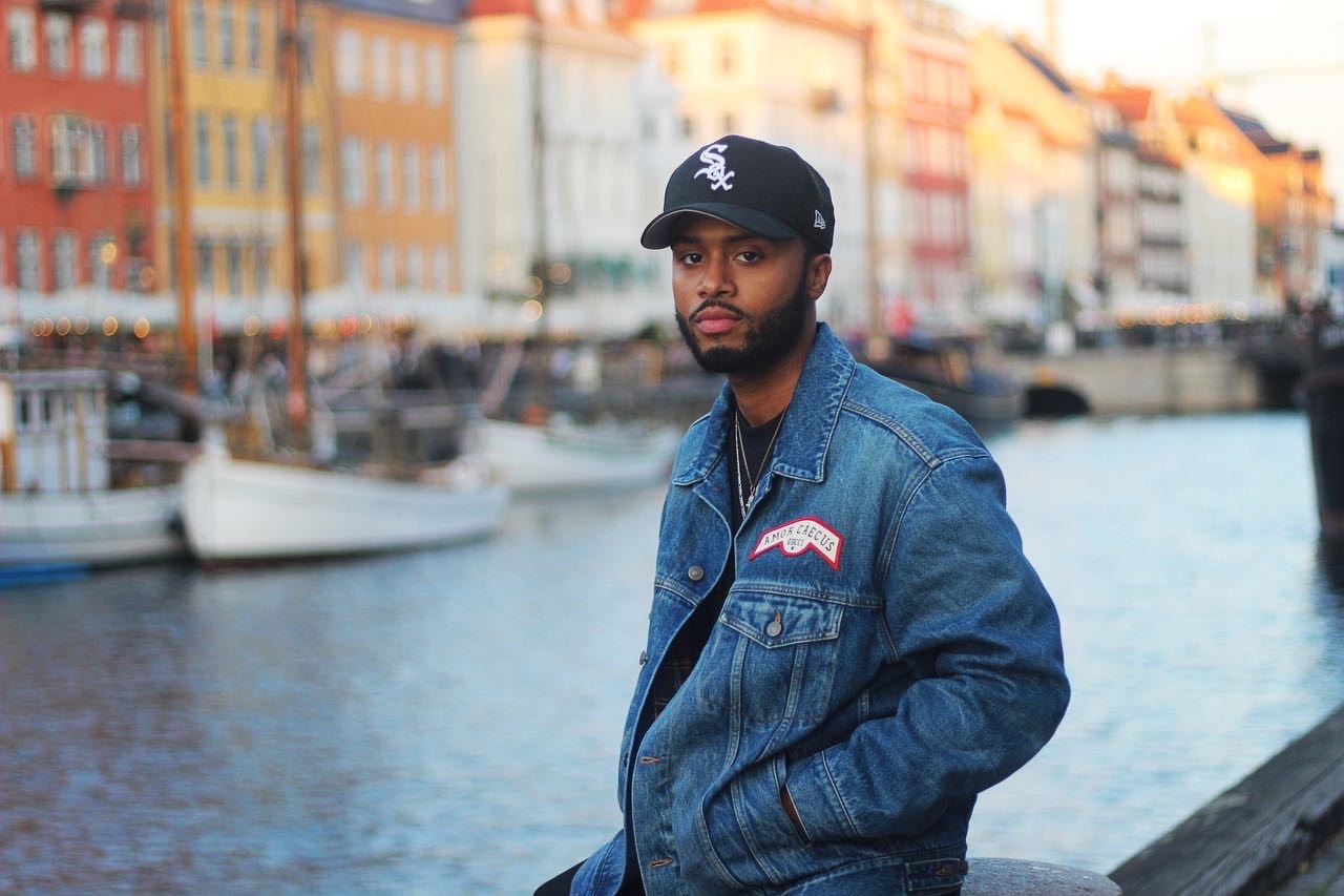 Mark wearing a denim jacket and a baseball cap posing for a portrait photo along a canal with colorful buildings in the background