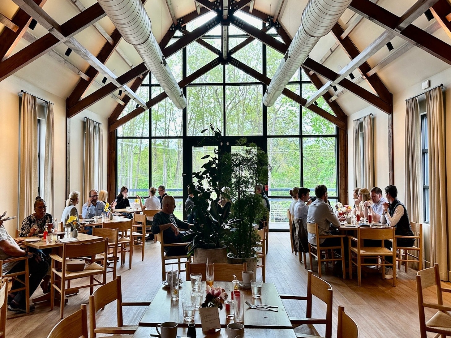 The image features a group enjoying a meal in a spacious dining area with high ceilings, expansive windows, and visible beams.