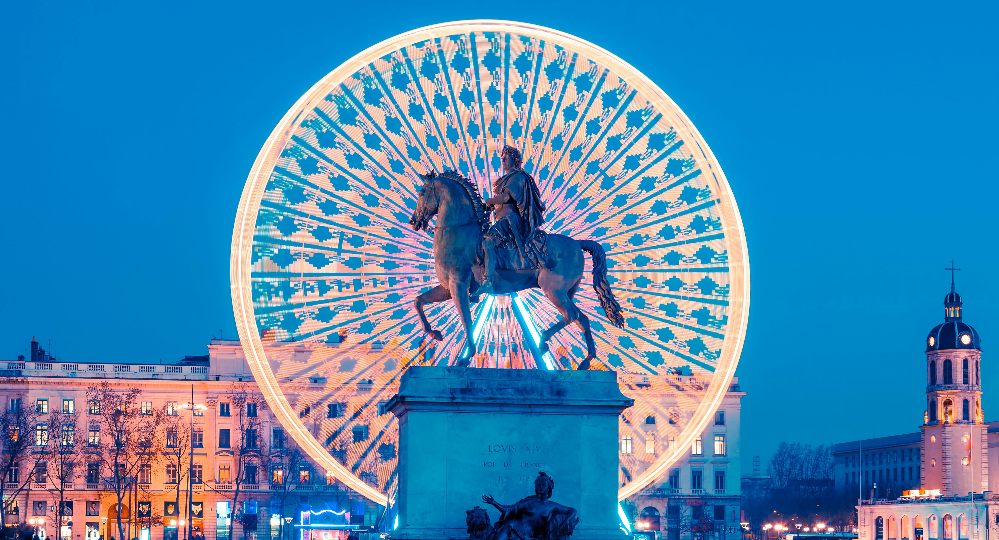 A monument of a man riding on a horse with city buildings in the background during the nighttime