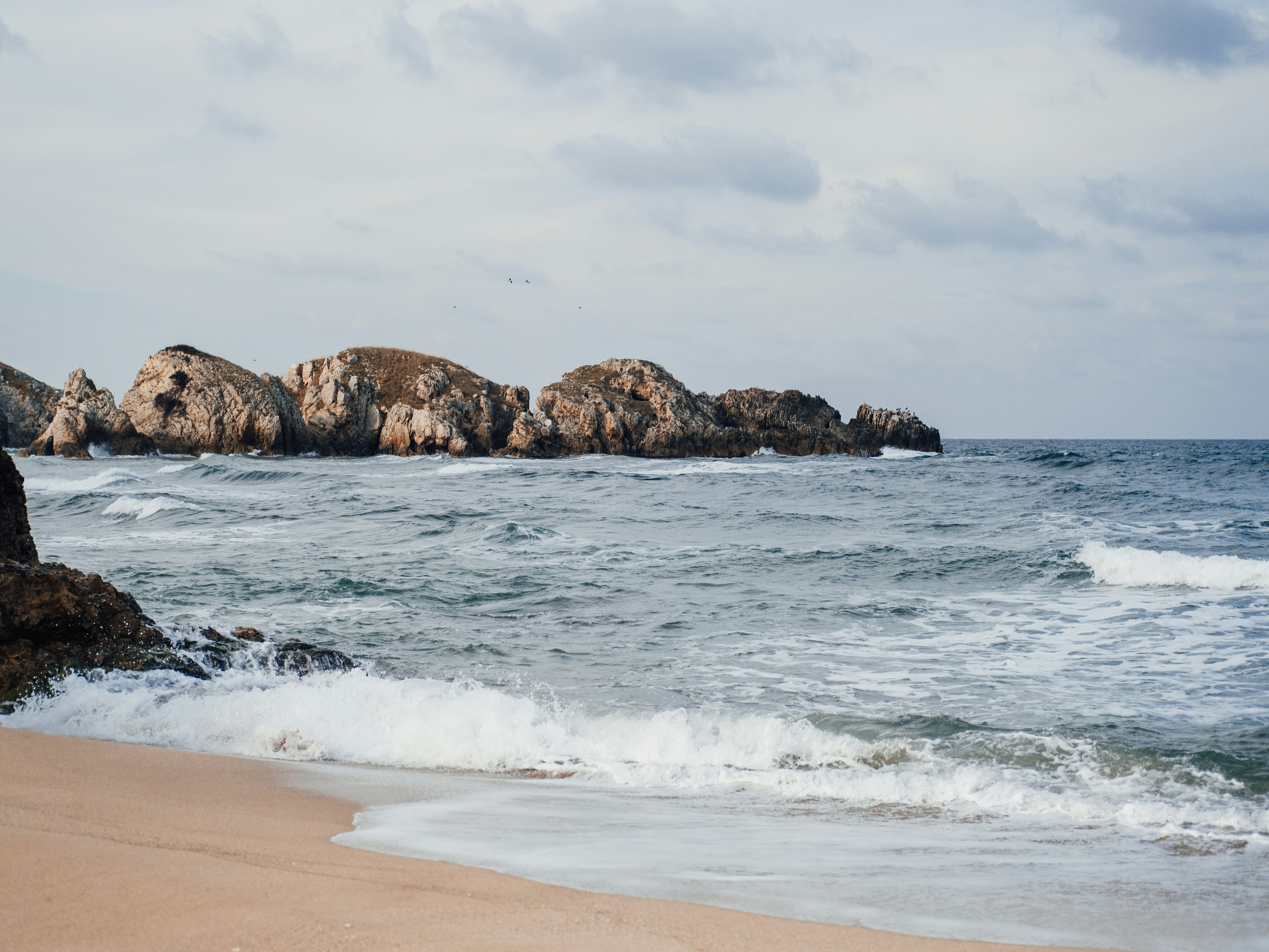 A tranquil beach scene with approaching waves and distant rocky outcrops under a cloudy sky.