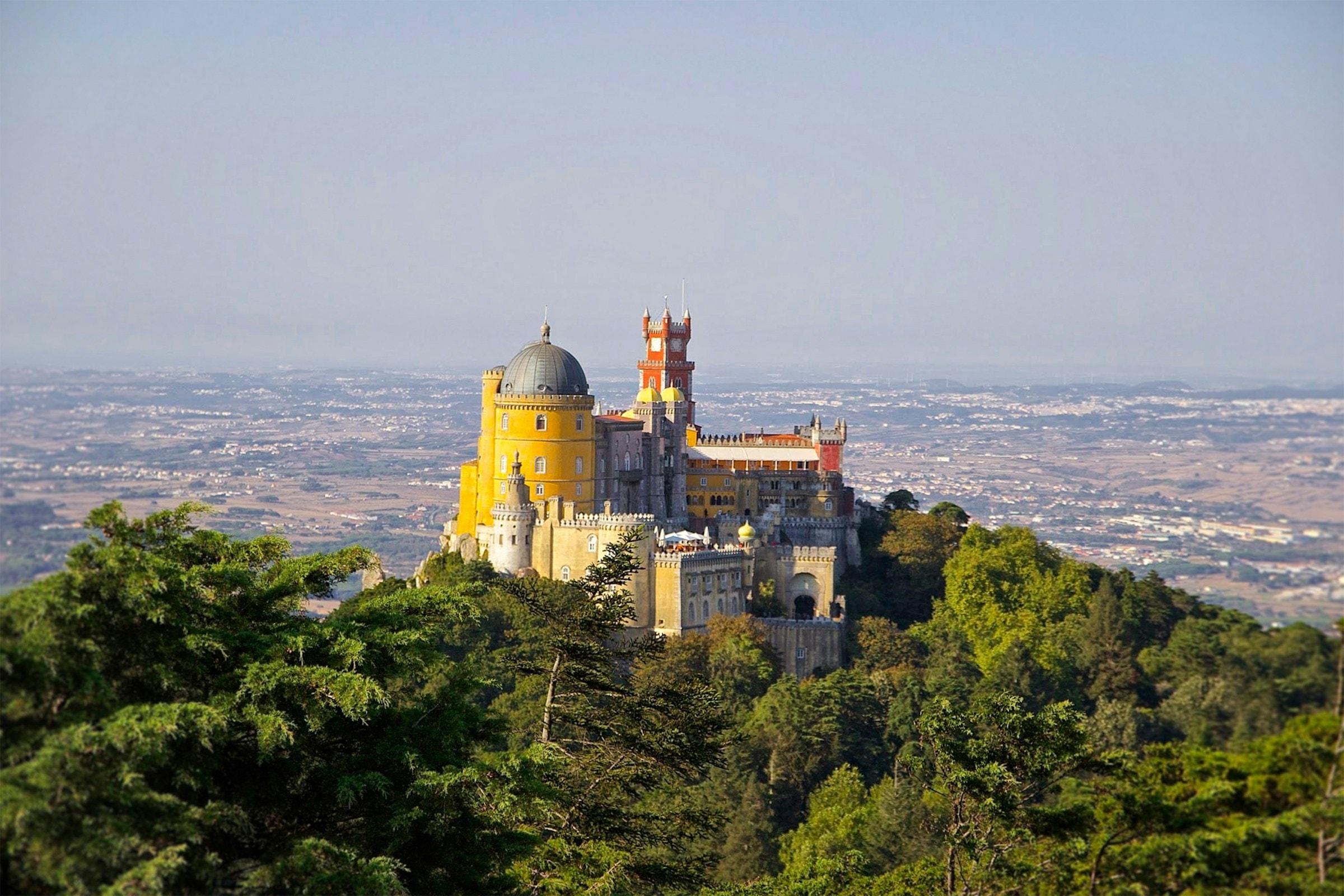 View of the golden and red, fairytale style Pena Palace of Sintra, sitting atop an emerald hill, surrounded by a forest, looking down at the landscape below.