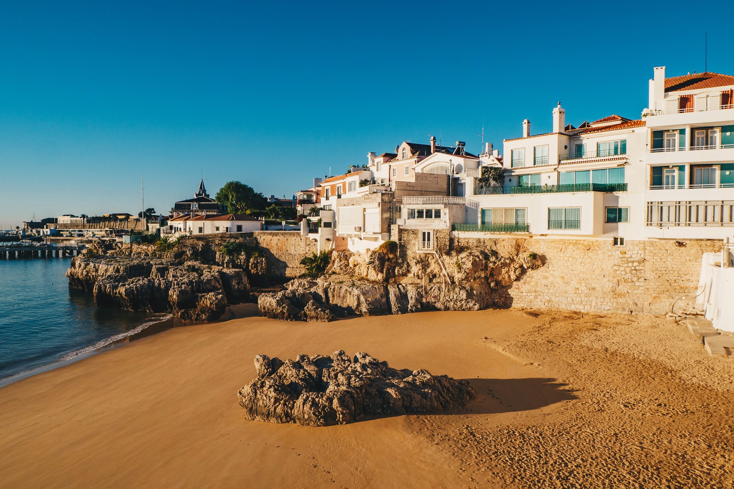 A small sandy area by the beach, with large rock formations and some residential buildings and hotels on a sunny day. 