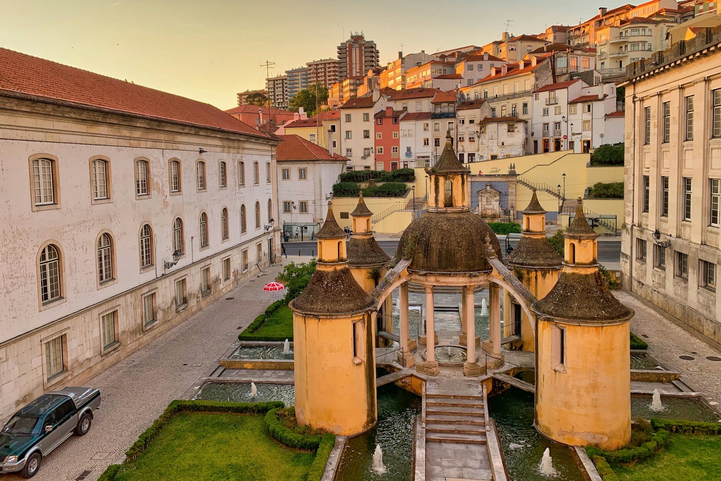 A courtyard with grass, water and circular shaped architectural structures surrounded by historic buildings of a town.