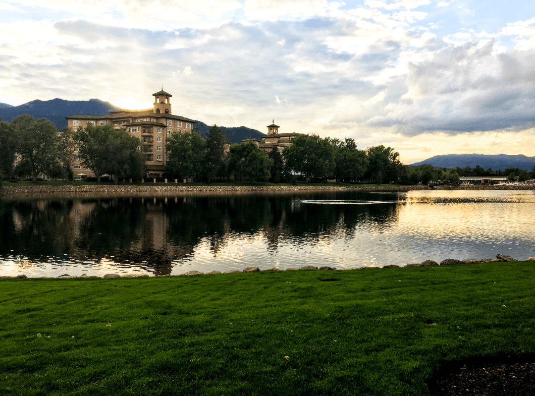 A view of green grass, a reflective pond and a beautiful building surrounded by trees and mountains in the background. There is a golden sunset reflecting over the water. 