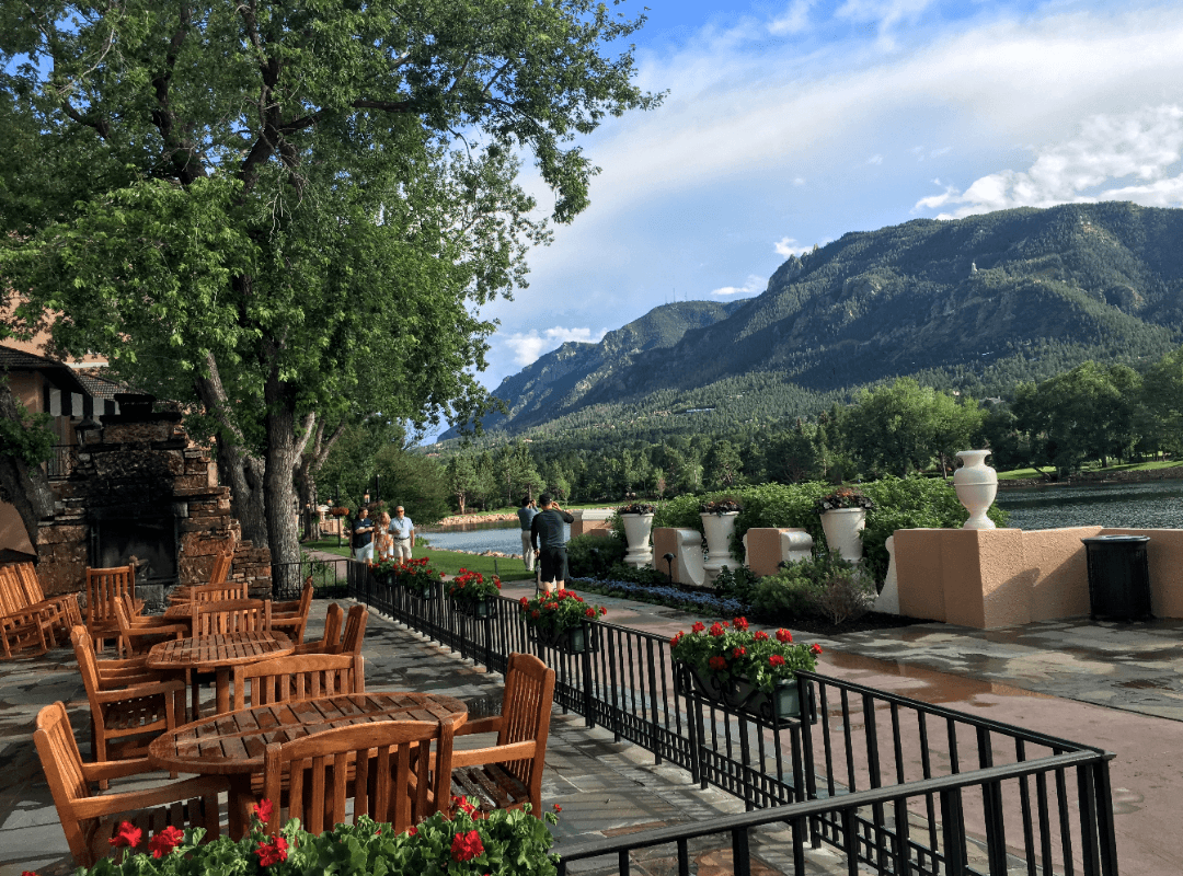 A picture of an outdoor patio with wooden tables and chairs, a sidewalk, planters, trees and a stunning view of a mountain range toward the right side of the image. 