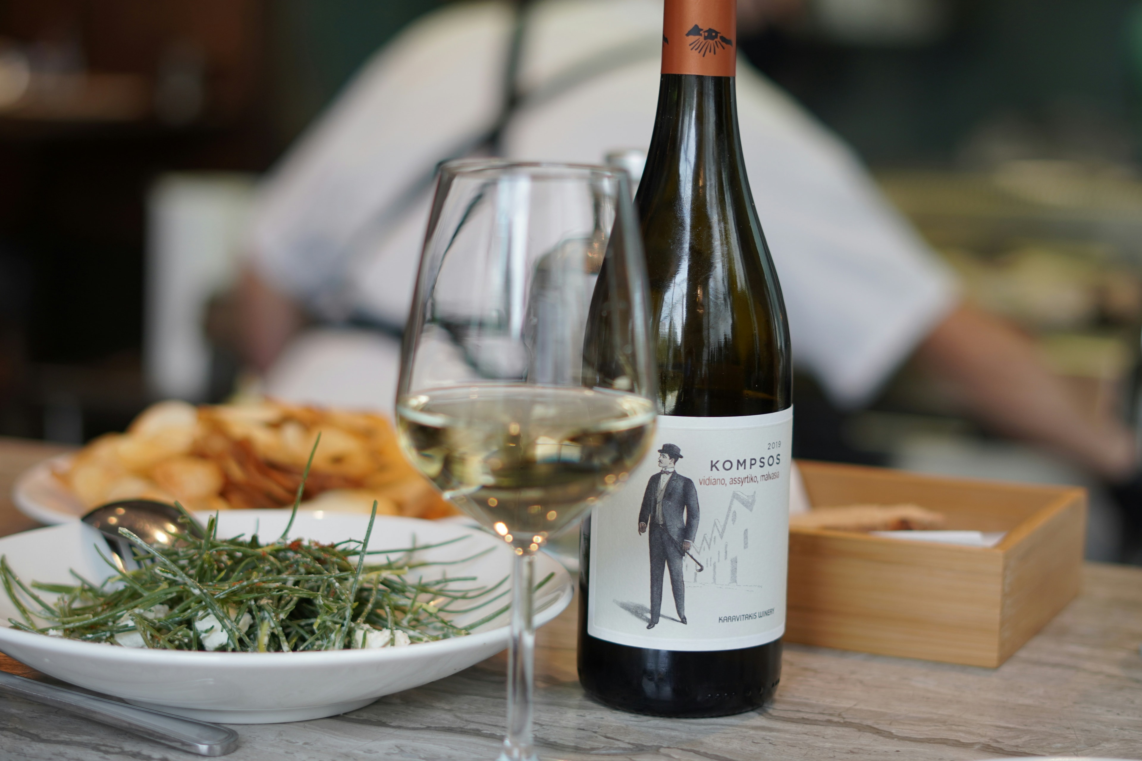 A wooden table with a wine bottle, glass of white wine and a white plate with green vegetables.