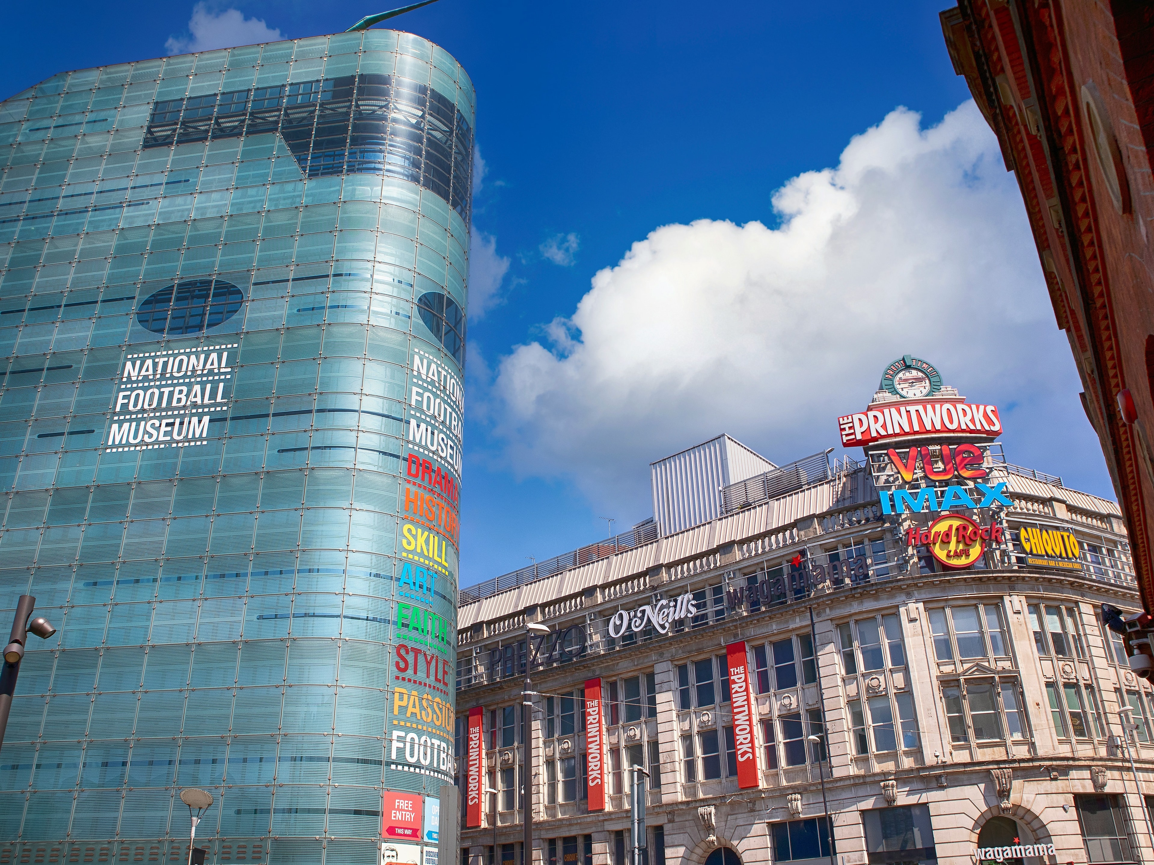 The image features the National Football Museum and the Printworks entertainment venue in Manchester under a clear blue sky.