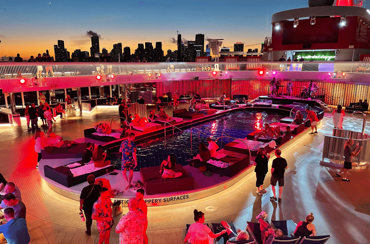 A view of the outdoor deck of the cruise at night with purple and pink lighting and people lounging by the pool in a party atmosphere