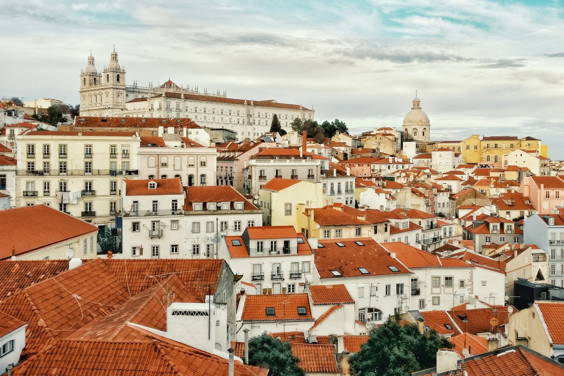 A view of Lisbon's white buildings, topped with red roofs and a domed church against a semi-cloudy sky.