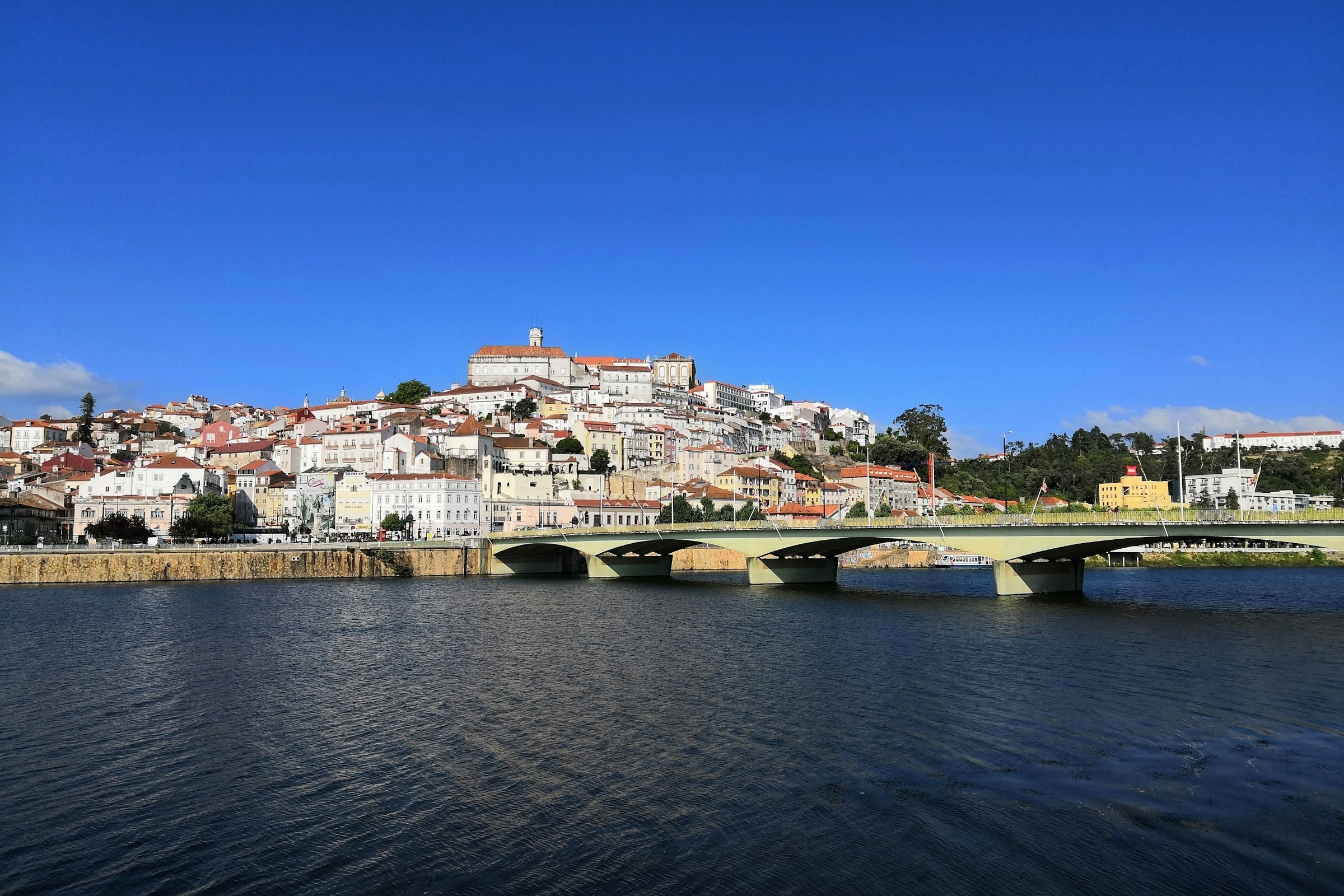 A view of pale-colored buildings from across a river, with a bridge on a sunny day.