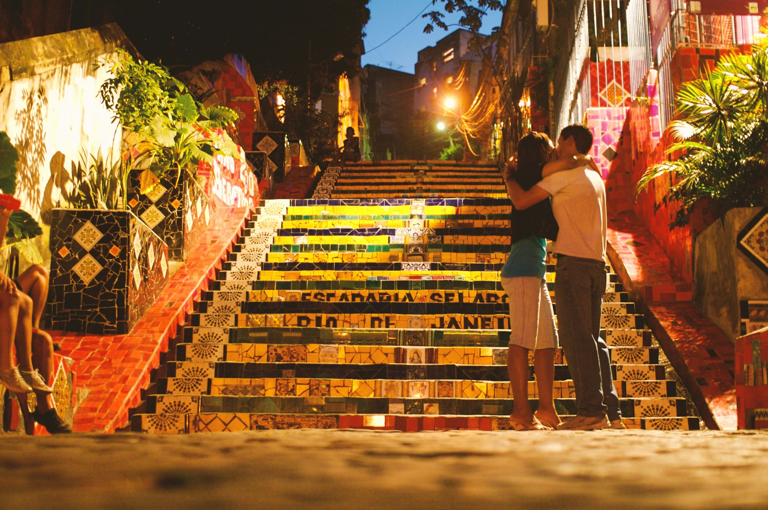 The image depicts the vibrant and colorful Selaron Steps in Rio de Janeiro, Brazil at night with a couple kissing