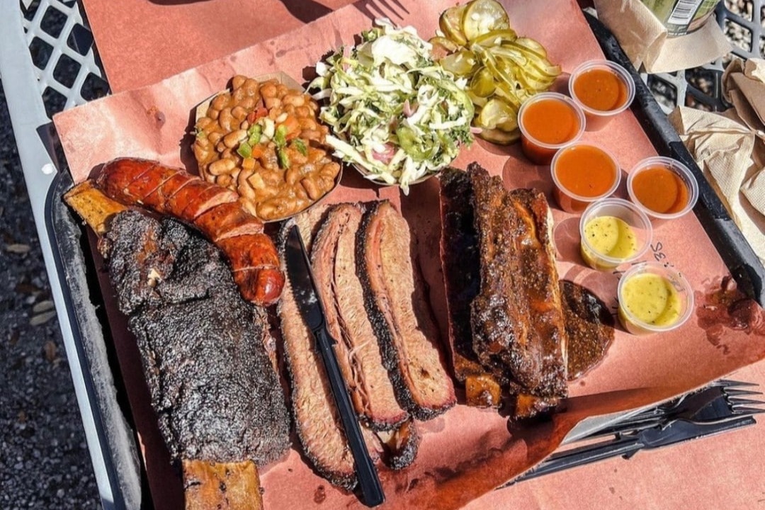 A platter of smoked meats and cups of various sides on a red tablecloth 