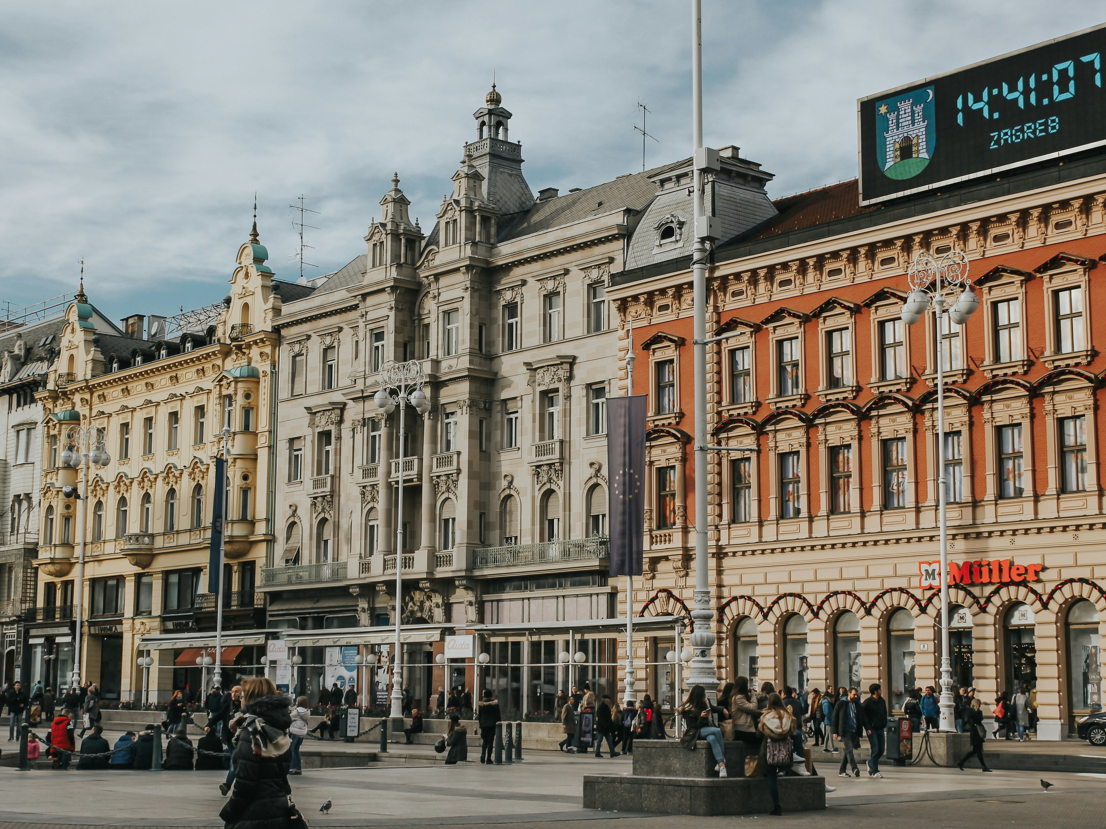 A bustling city square with historic architecture and a digital clock displaying time and temperature.