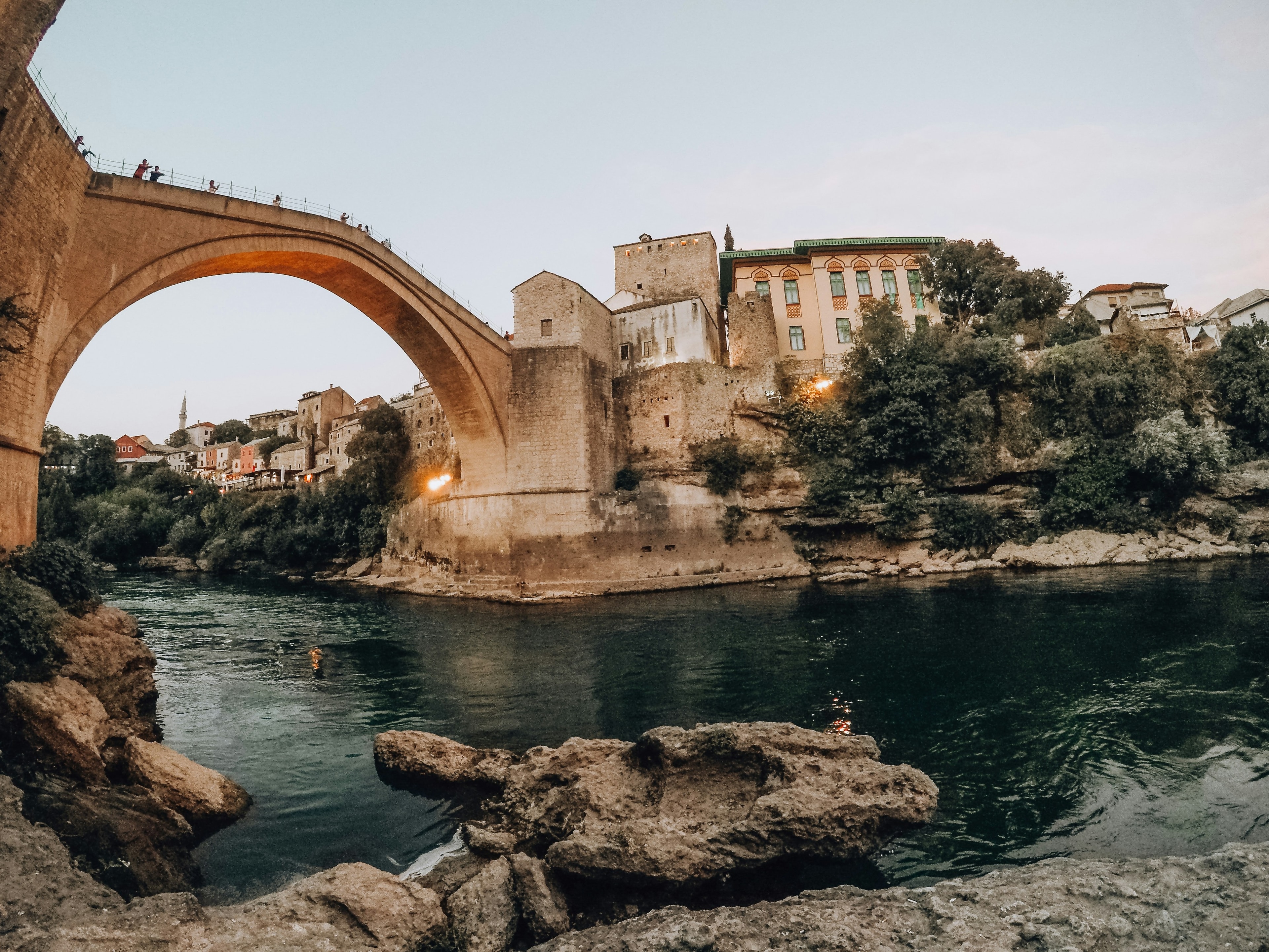 An old stone bridge arching over a river, with buildings on the banks under a dusk sky.