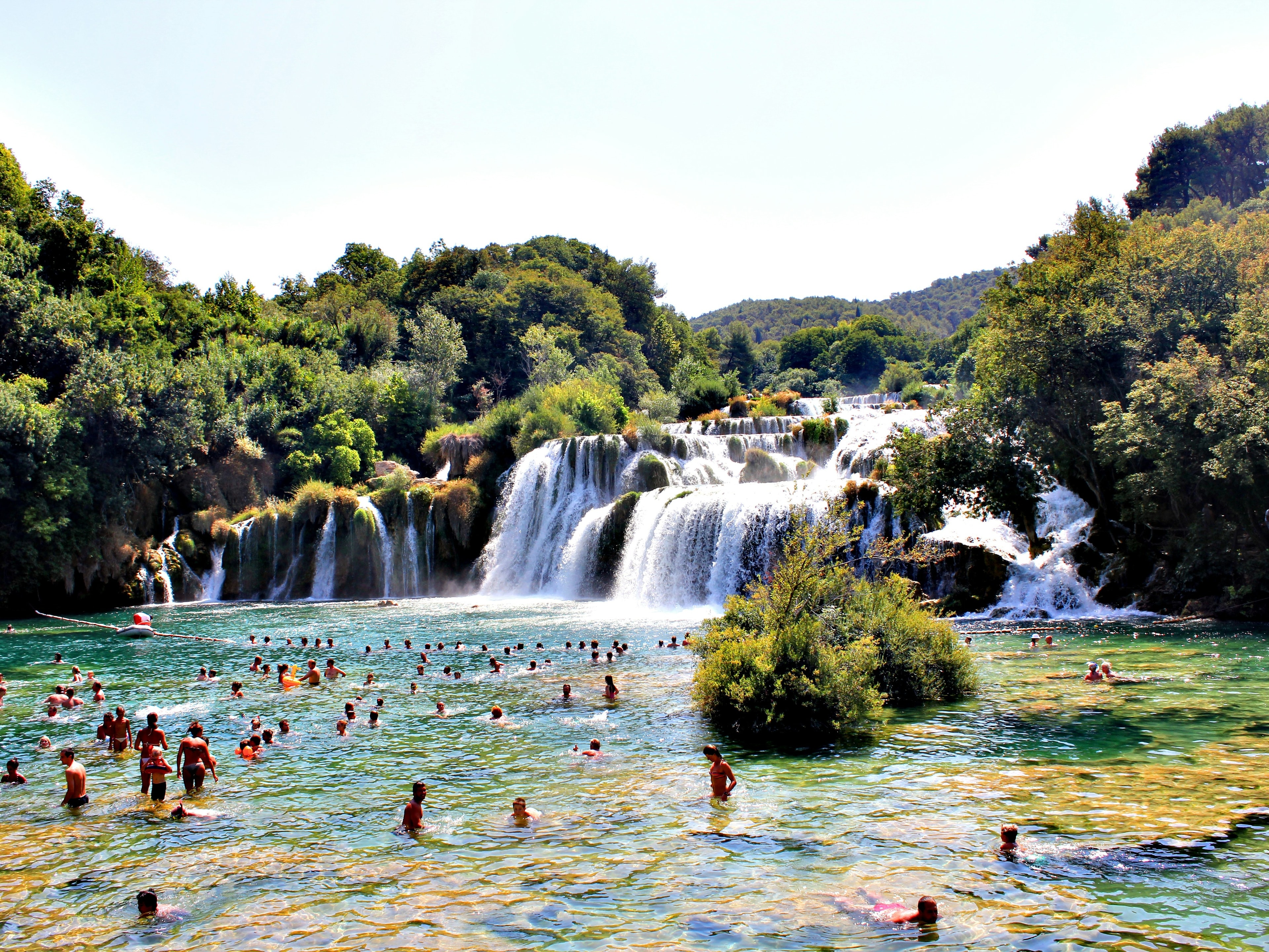 A scenic swimming spot with people and cascading waterfalls amid lush greenery.