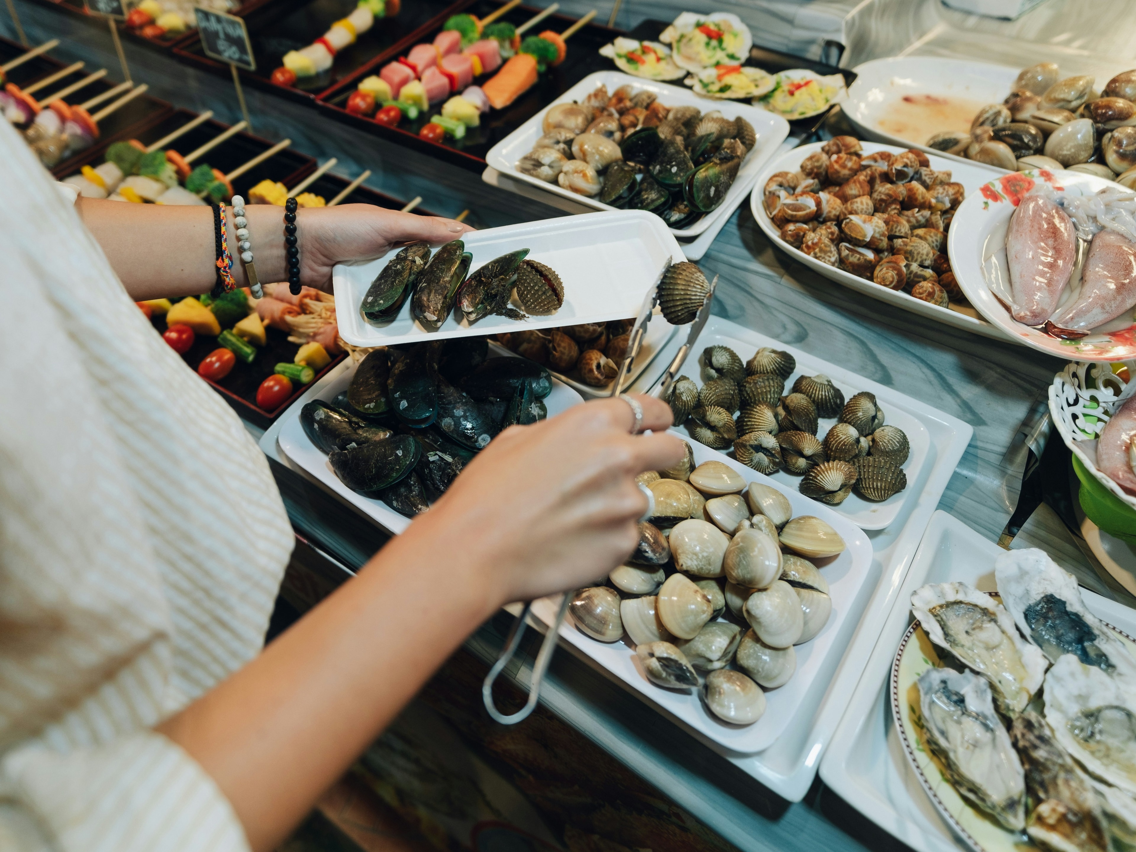 A buffet selection featuring a variety of seafood dishes, with a person picking shellfish using tongs. 