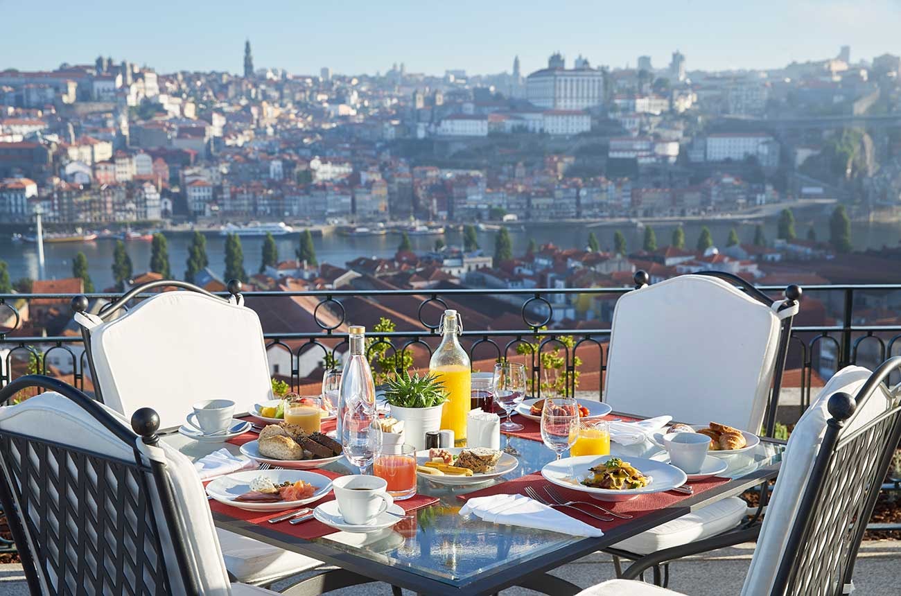 A view of a table on an outdoor terrace, set with different plates of food, and bottles of water and orange juice, overlooking the city.