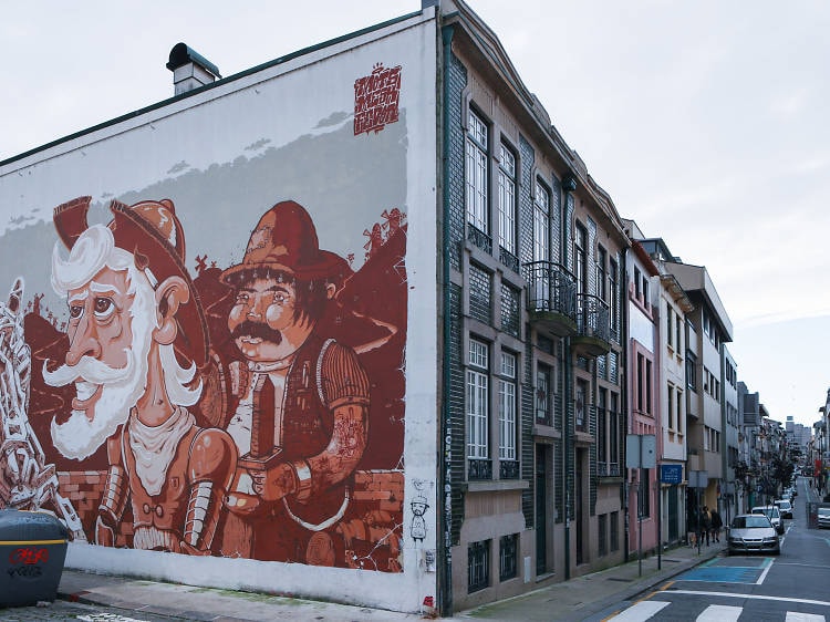 A massive mural on the exterior wall of a building in  Porto, with a view of the street and parked cars. 