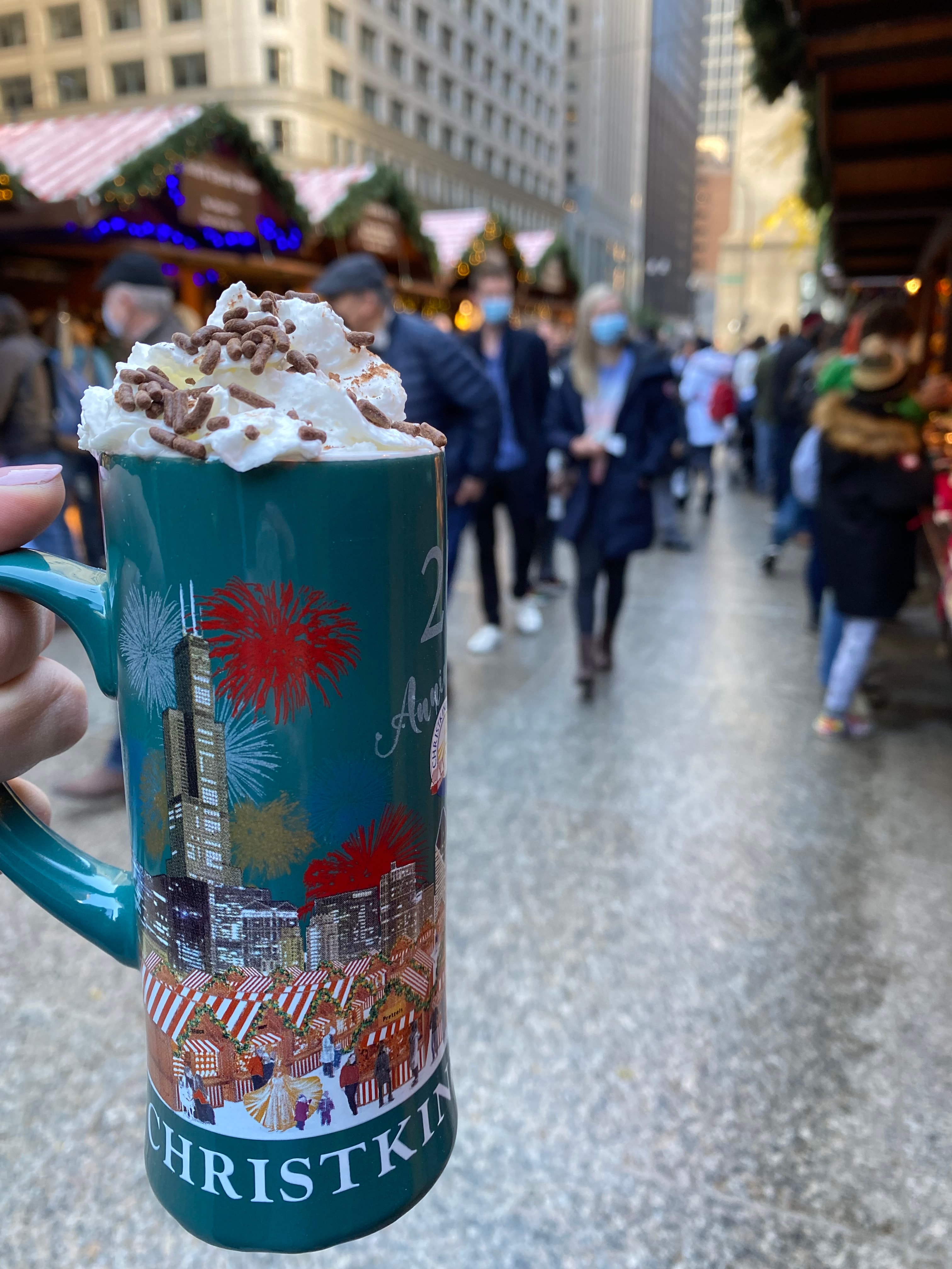 A large close-up shot of a festive mug with a warm Christmasy-drink inside, topped with whipped cream and chocolate sprinkles, with a street scene in the background. 