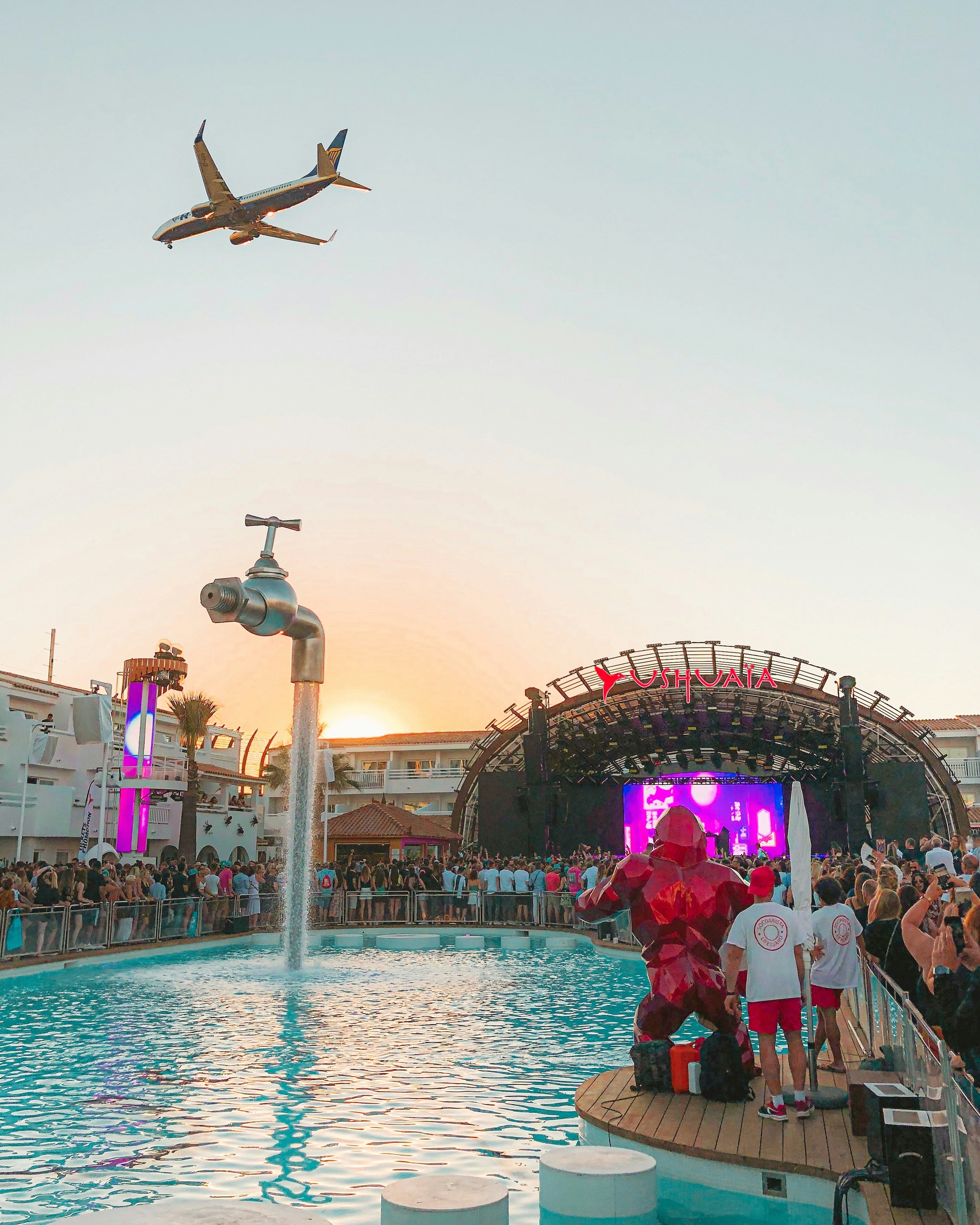 A giant pool party in Ibiza, with a stage, many people and an airplane flying above the festivities.