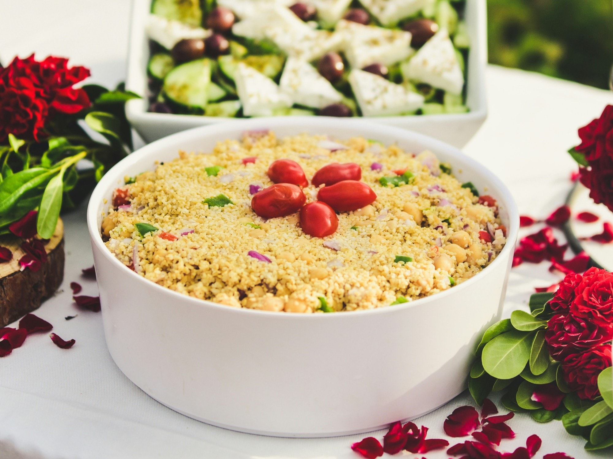 A table set with a massive bow of cous-cous, topped with cherry tomatoes, a bowl of feta cheese, cucumbers and olives and red flowers.