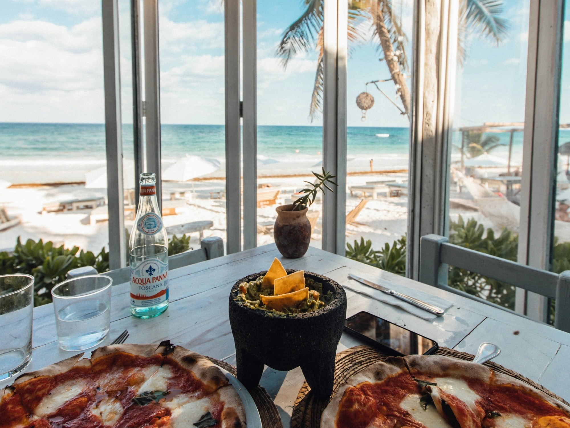 A view of a table at a restaurant adorned with two pizzas, and a clay bowl of guacamole, with a view of the beach and sea through the open windows.