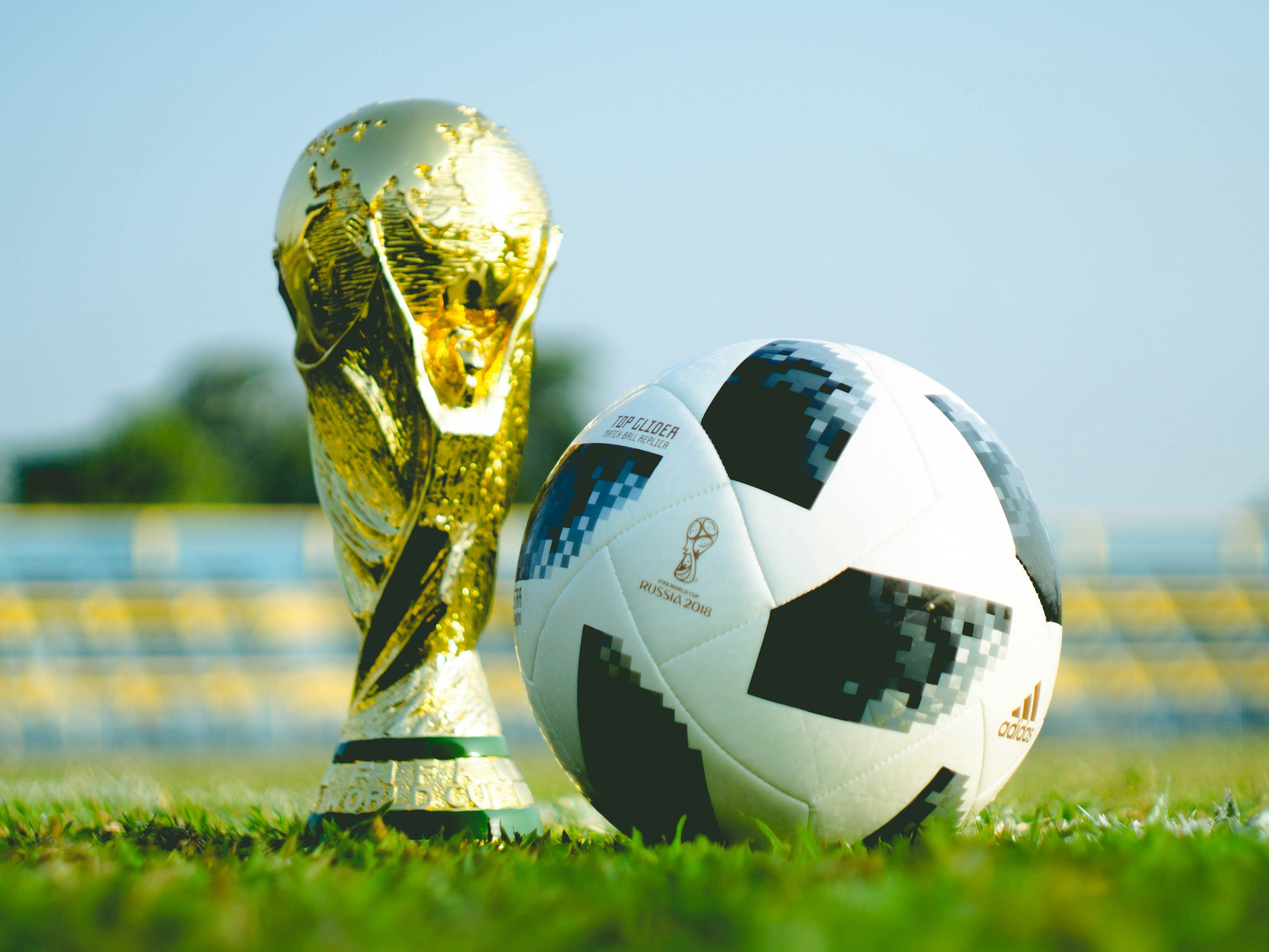 The image features a trophy and a soccer ball on a grass field, with a backdrop of empty stadium seats, evoking the anticipation of a grand sporting event. 