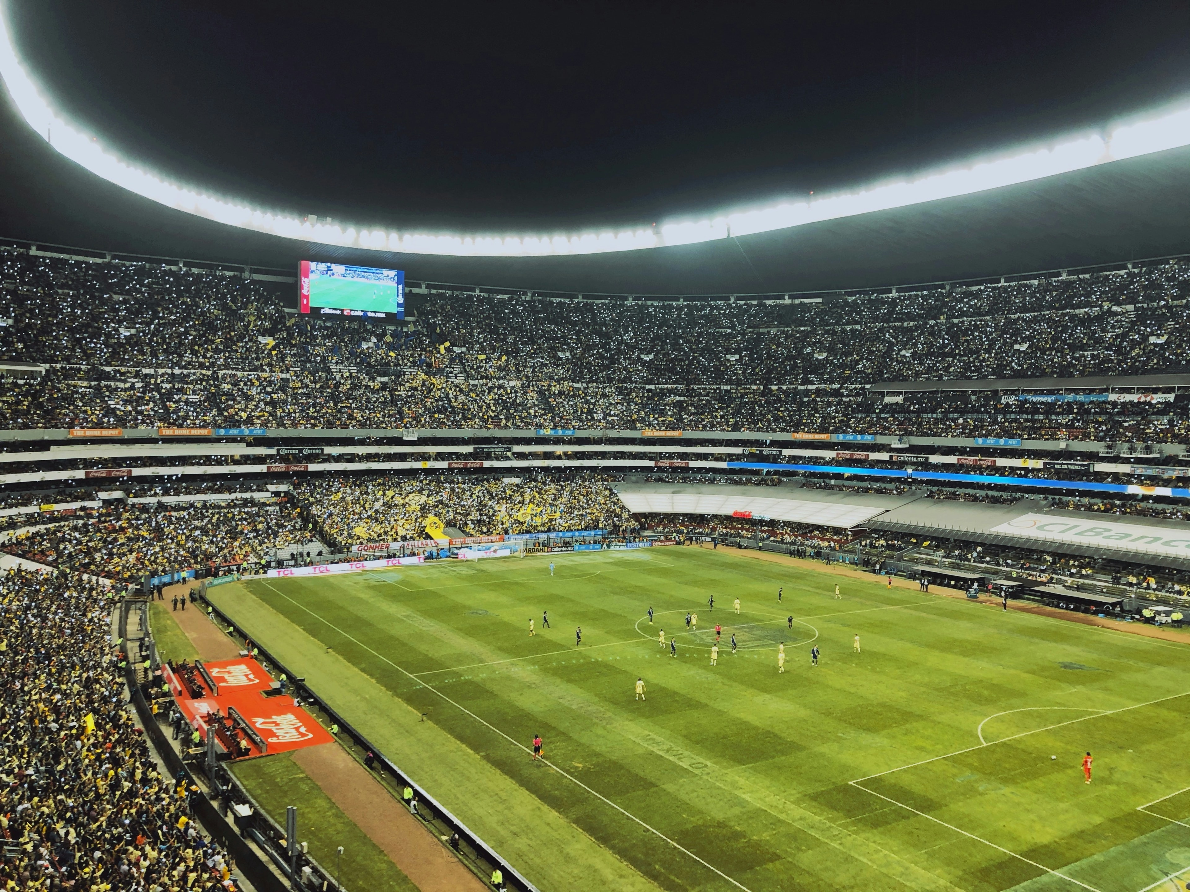 The image showcases a vibrant soccer stadium bustling with fans and action during a nighttime match, under a luminous closed roof.