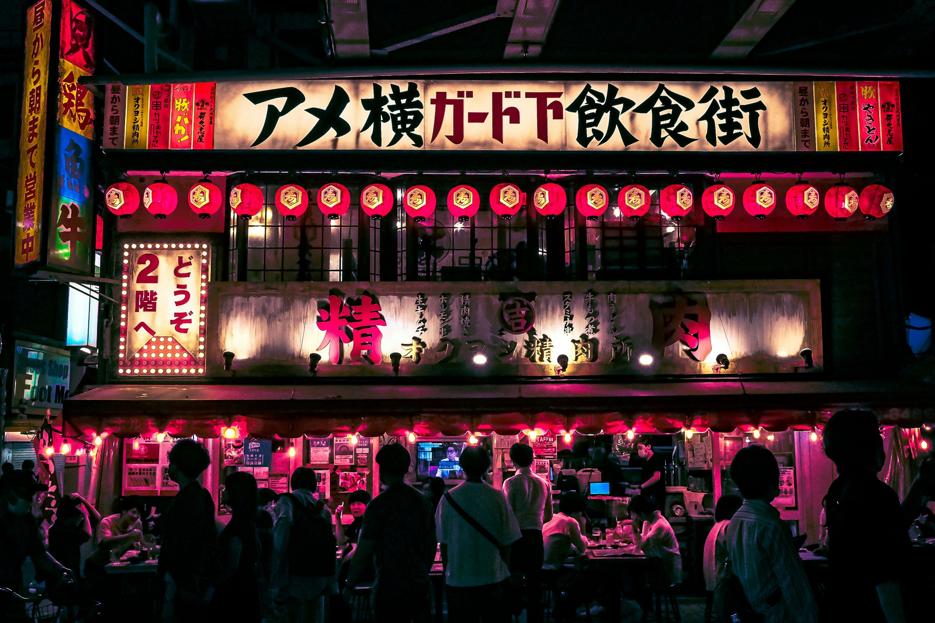 People in front of colorful illuminated signs and red lanterns at a bustling restaurant.