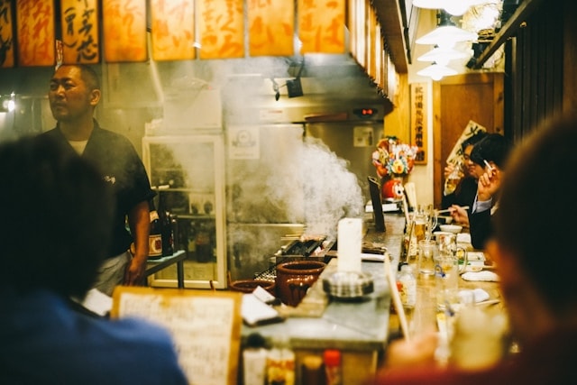 An inside peak of a traditional Japanese with an open kitchen, a chef and patrons sitting at the counter enjoying their meals.