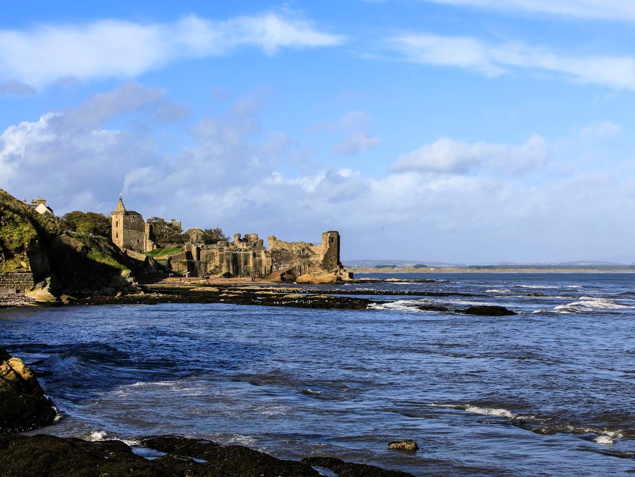 A view of the rippling blue sea in Scotland, with stone castles and rolling green hills in the background. 