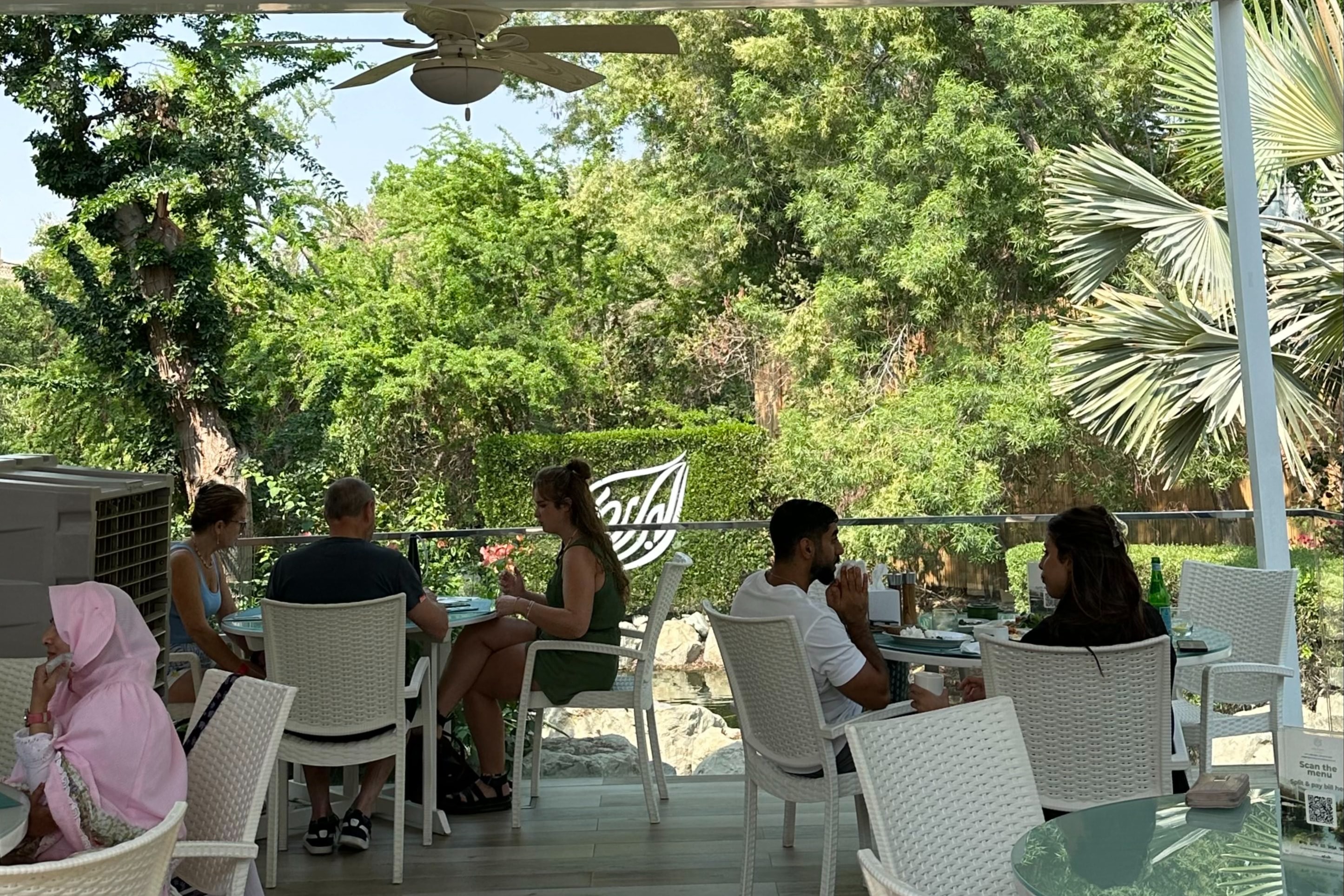 A patio full of white chairs and tables with people seated and dining. There is a lush, green forest full of trees in the background. 