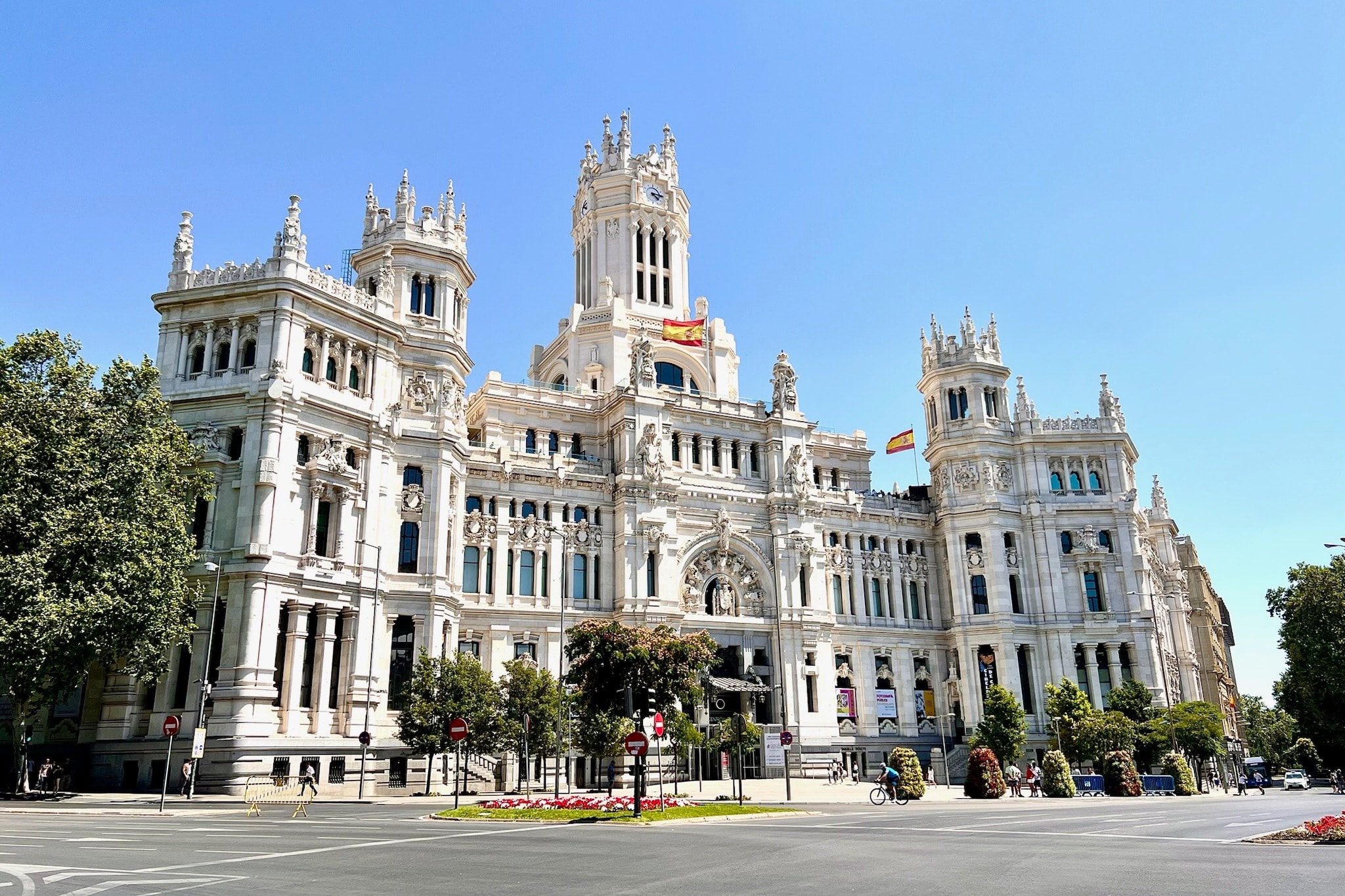 A large white building with the Spanish flag and beautiful detailing. There are trees and bushes in front of it and a blue sky in the background. 