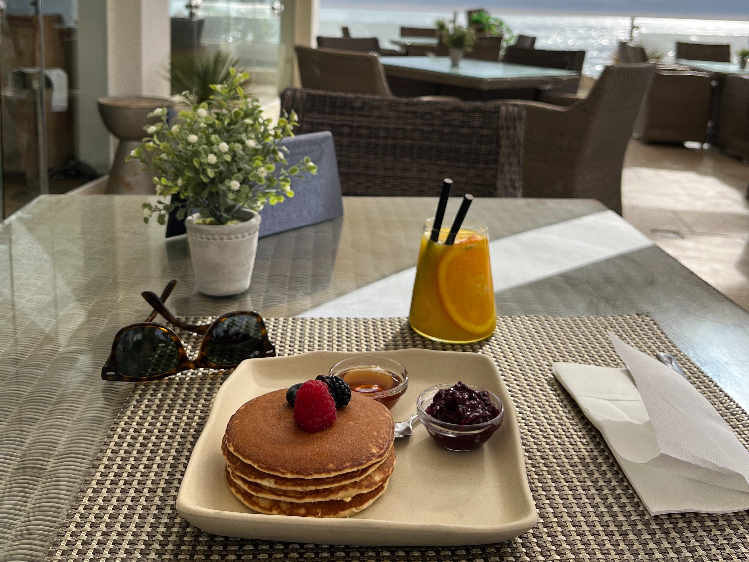 A glass table with a square plate full of pancakes and a bowl of jam. There is a drink with an orange slice and two black straws in the upper right hand corner and black sunglasses placed in front of a potted plant on top of the table. 