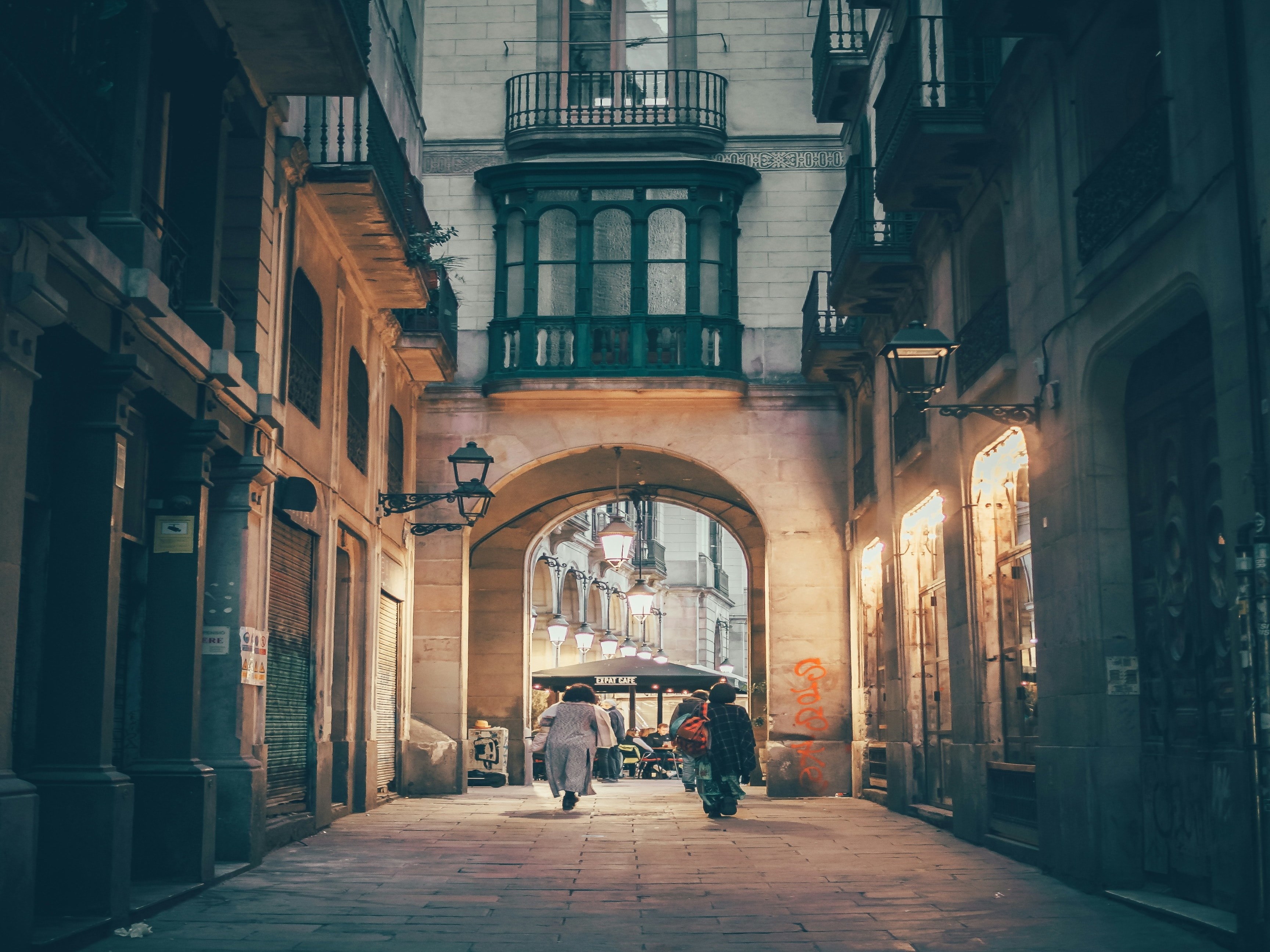 An old street alleyway with stone buildings on the left and right sides. There is a stone archway in the middle front of the image with people walking beneath it. You can also view metal balconies in front of the windows on the building. 