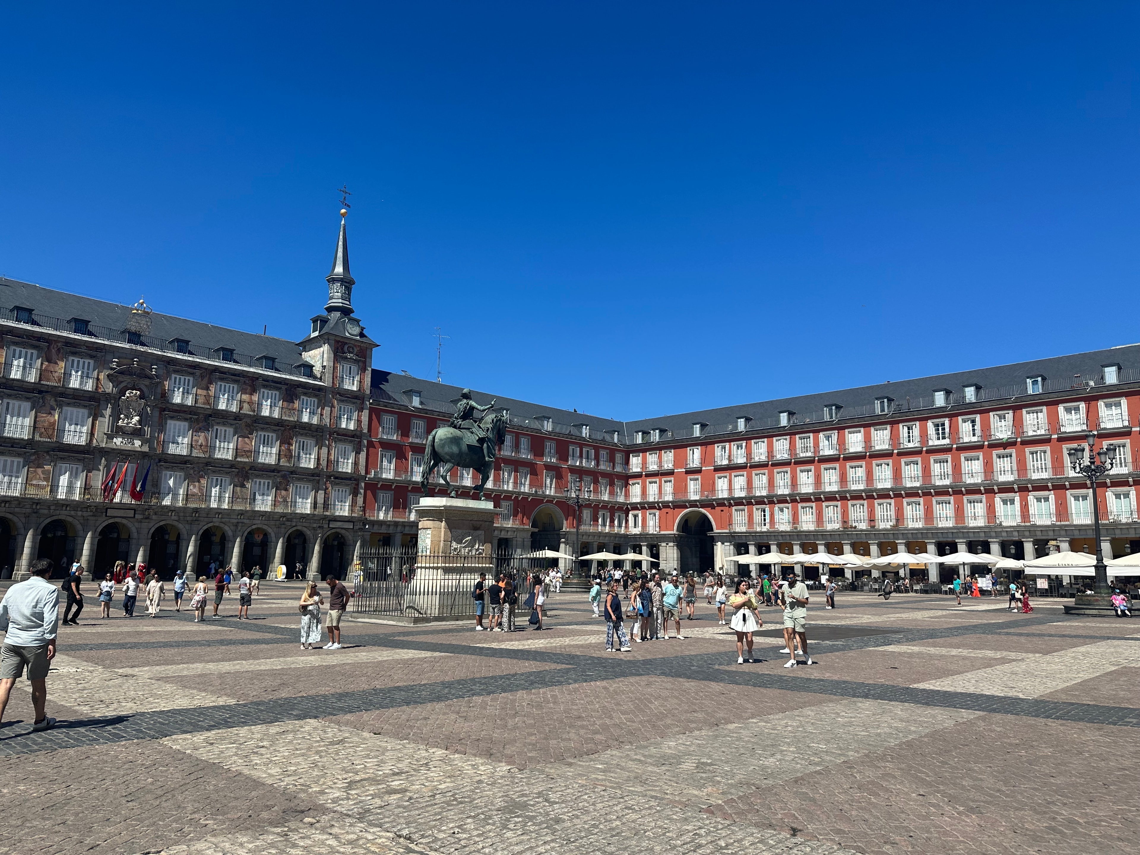 A town plaza with a large statue in the middle, people walking around and a building with red detailing and a tower in the background. 