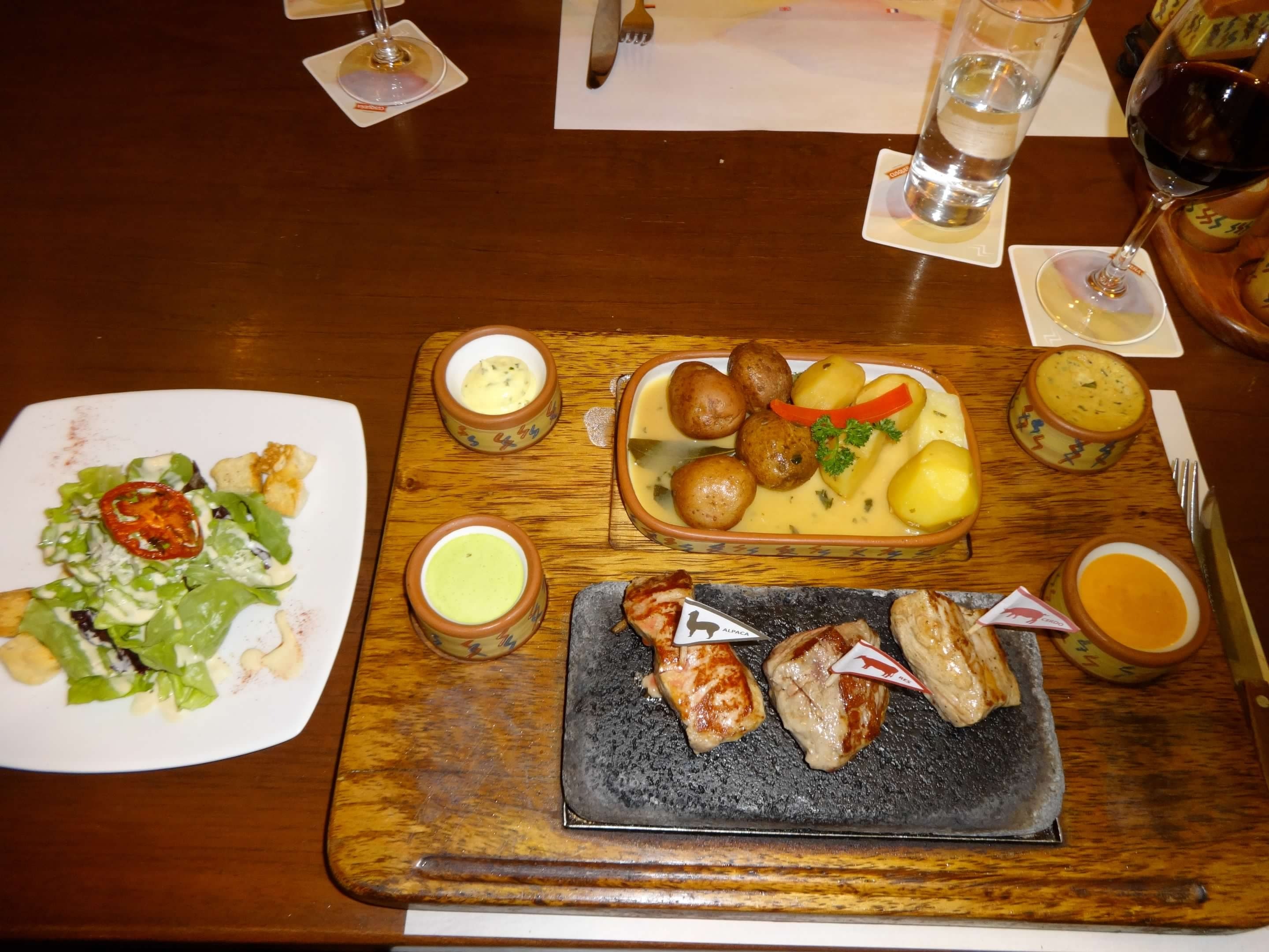 A picture of a wooden platter with various foods and sauces, next to a white plate with a dinner salad on top of it. There is a glass of water in the background next to a glass of wine, too. All of it is placed on top of a wooden table. 