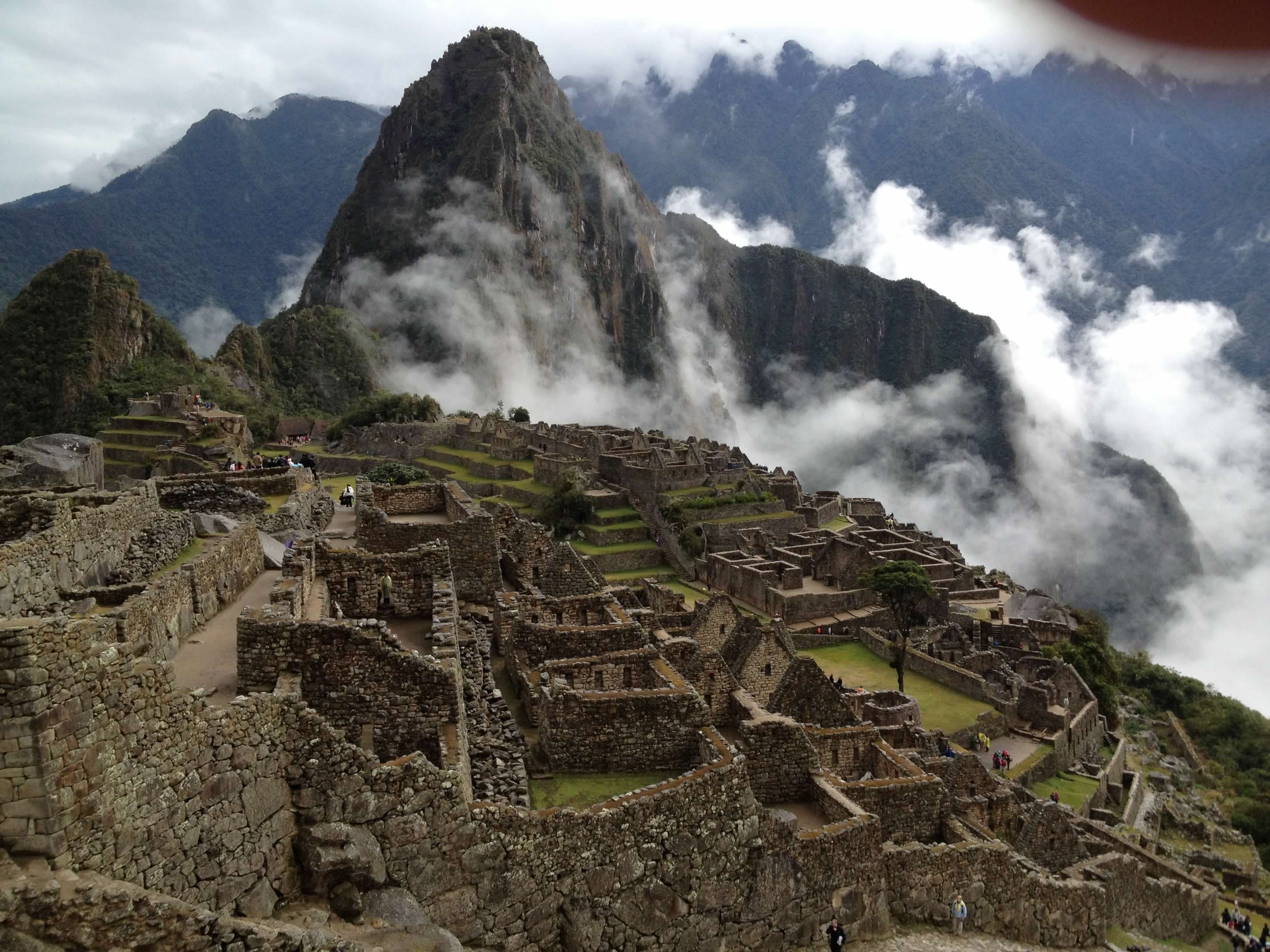 A stunning view of Machu Picchu during daytime with it's stone walls and green grass. There are clouds swarming the mountain peaks.  