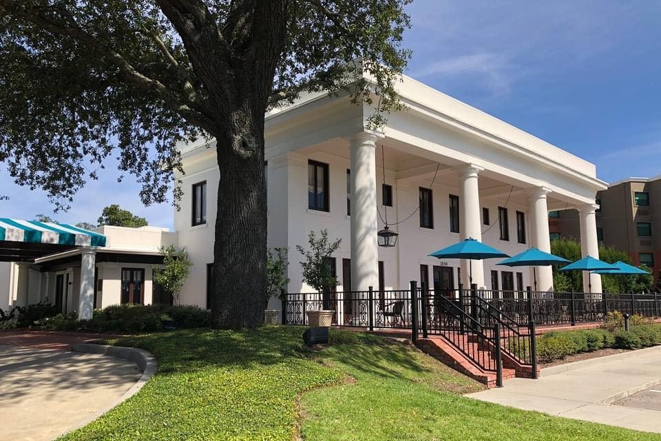 A large white home with porch columns and a large tree and blue umbrellas out front.