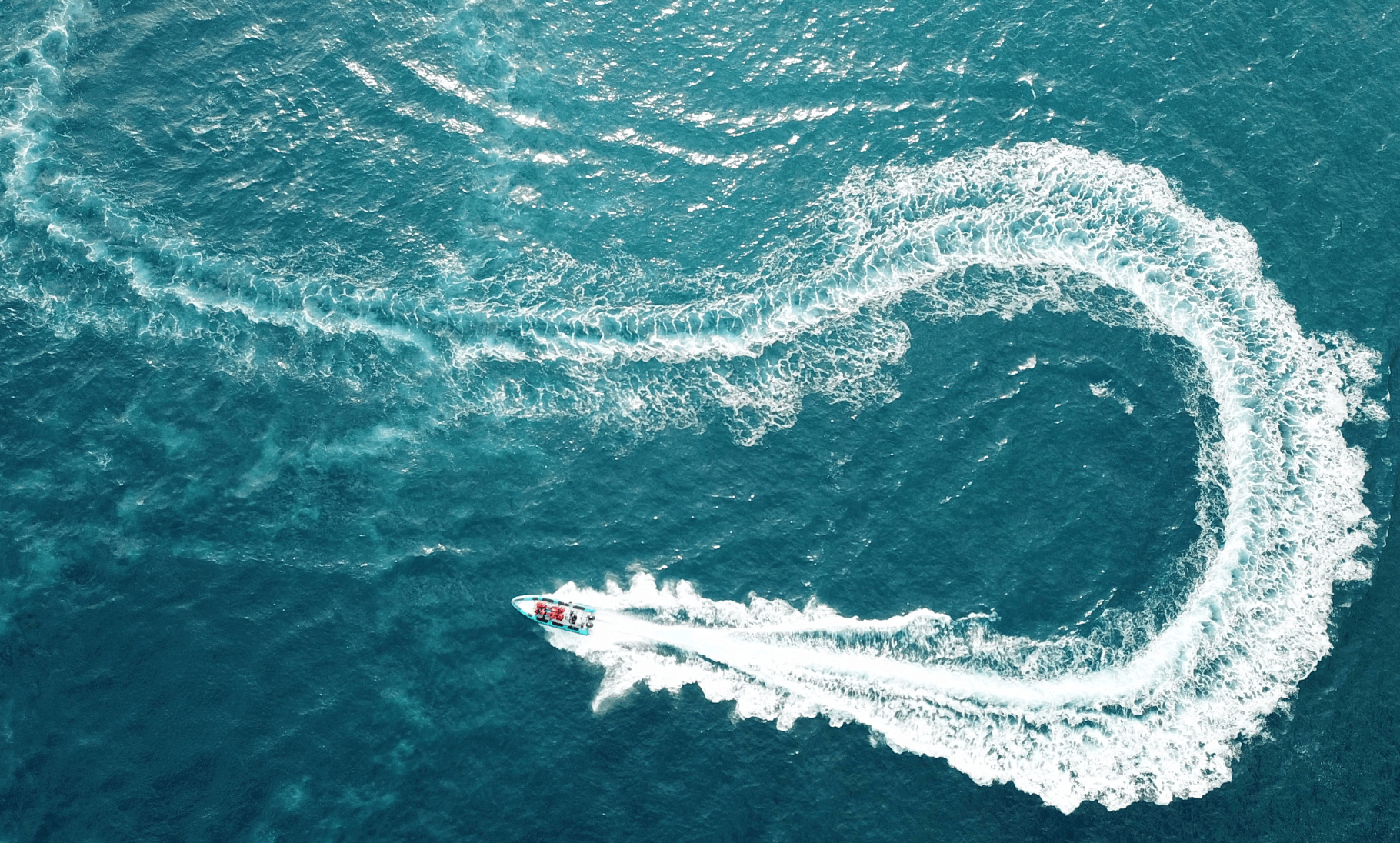 An aerial view of a yacht with curved waves over a turquoise blue body of water. 