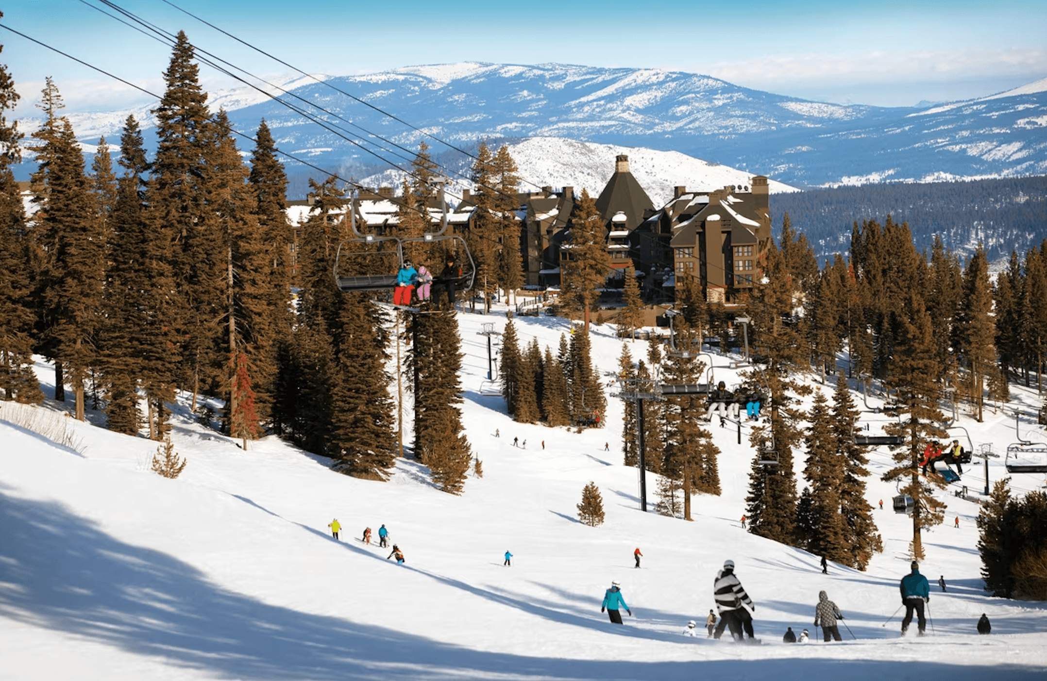 A beautiful ski hill surrounded by lush, green pine trees and various people in colorful ski gear on the snowy hill. There is also a snowy covered mountain in the far distance. 