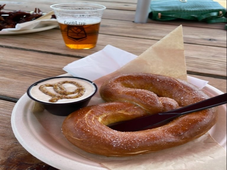 A pretzel with a side of mustard and plastic cup of beer served on a wooden table.