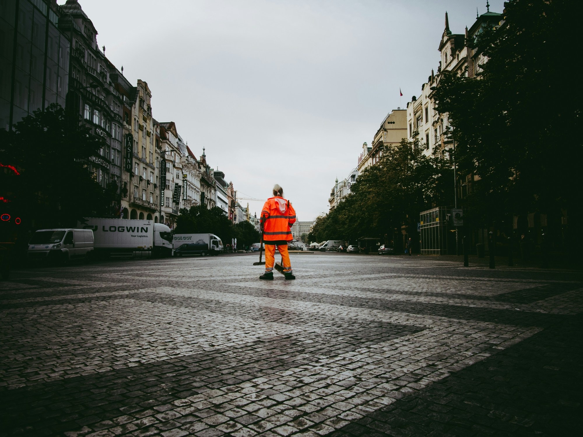 A brick road with a man wearing an orange outfit standing in the middle surrounded by stone buildings, houses and trees. 