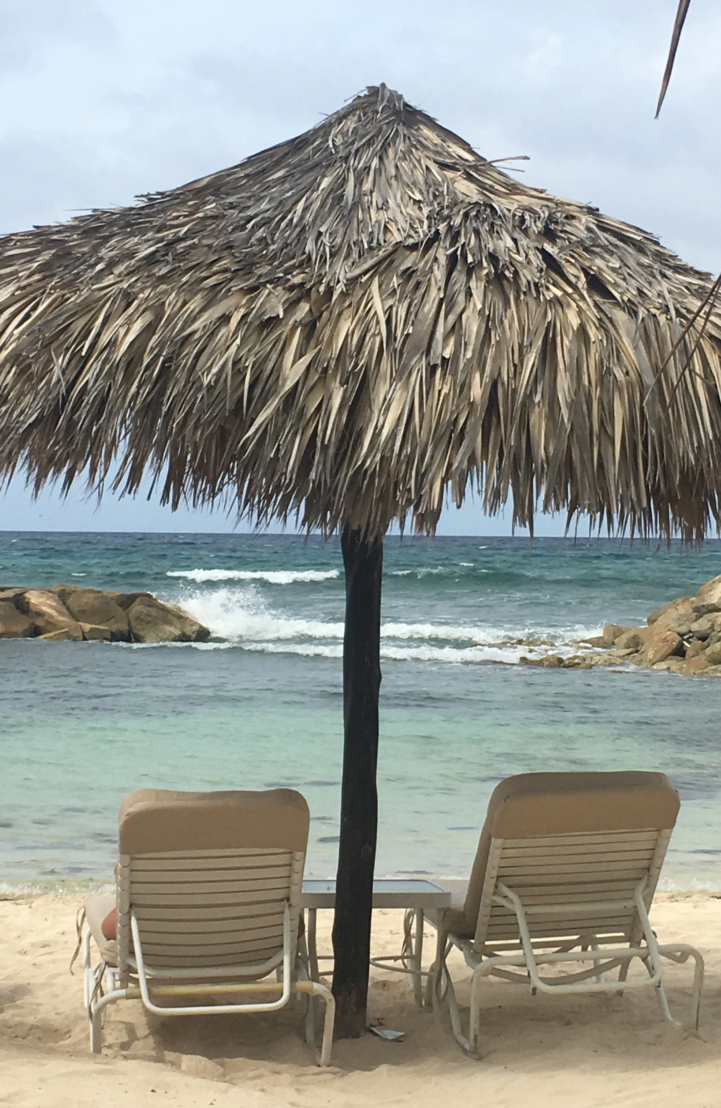 Two beach chairs under a straw umbrella looking out to the blue water and waves rolling in.